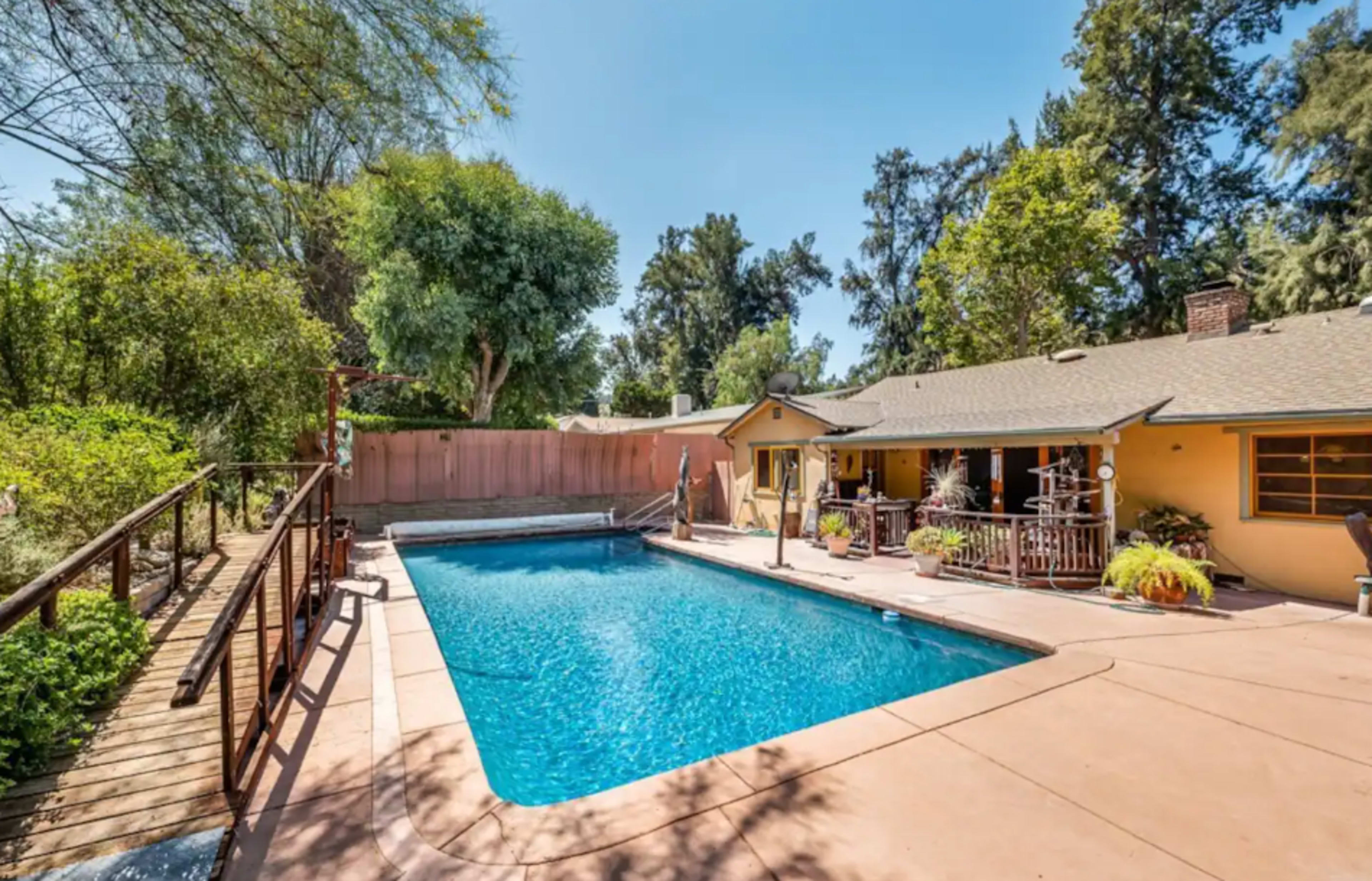 A swimming pool surrounded by a patio area and a house with a deck in a landscaped yard.