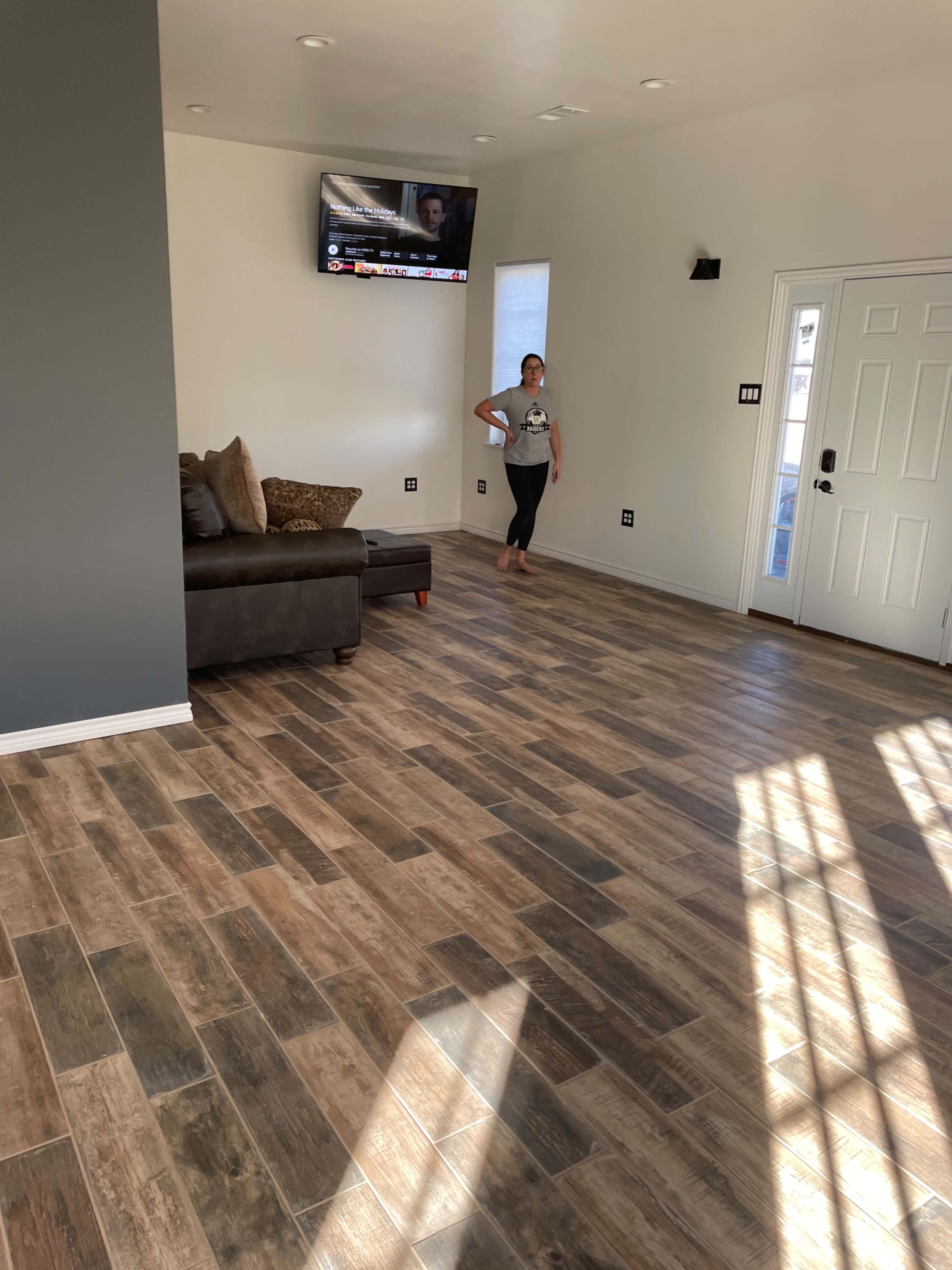 A woman stands barefoot in a spacious, well-lit living room with a dark sofa and a television mounted on the wall.