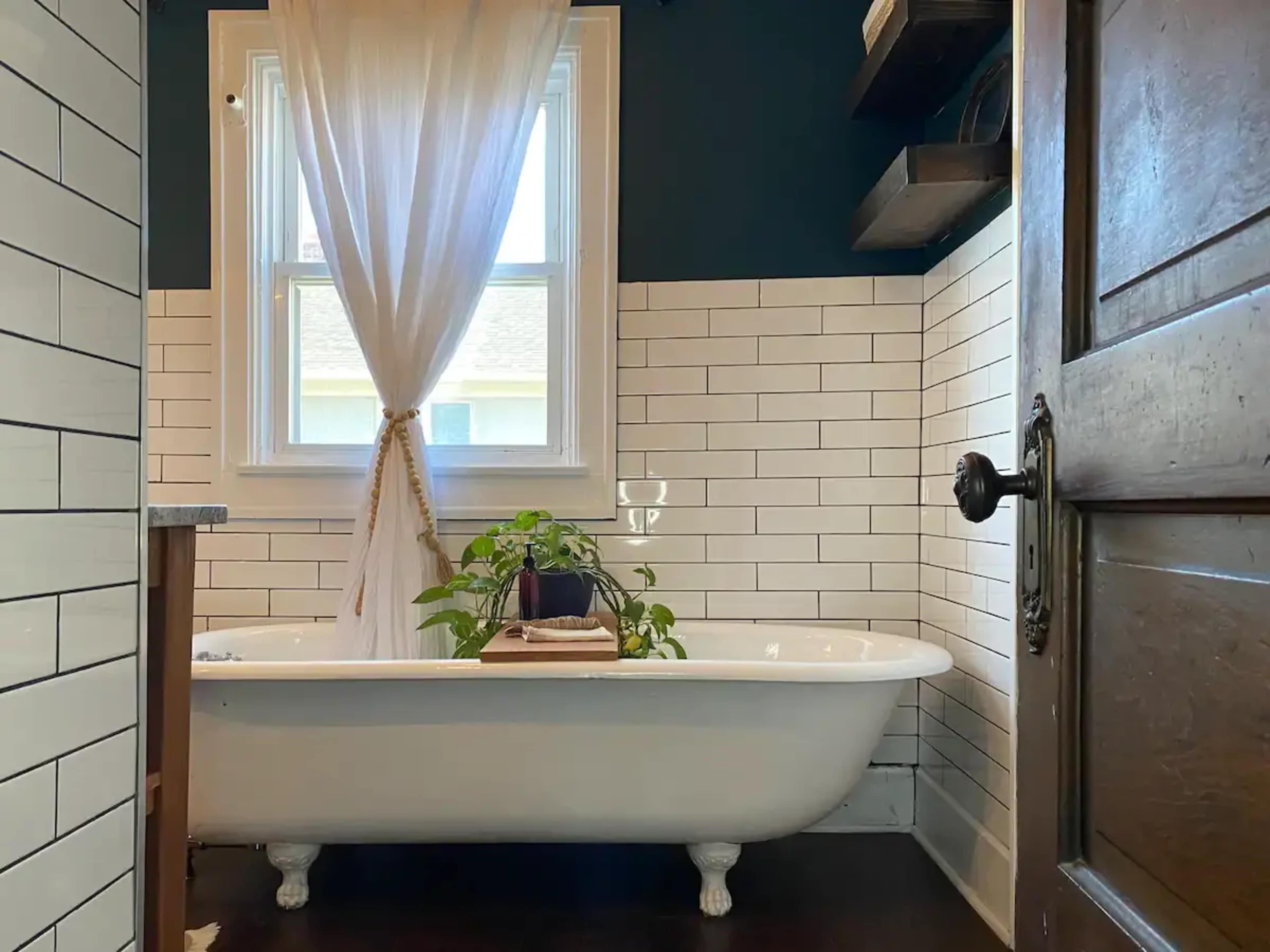 A clawfoot bathtub is positioned in a bathroom with white subway tile walls and a dark wooden door, featuring a plant and a glass of water on a wooden tray beside it.