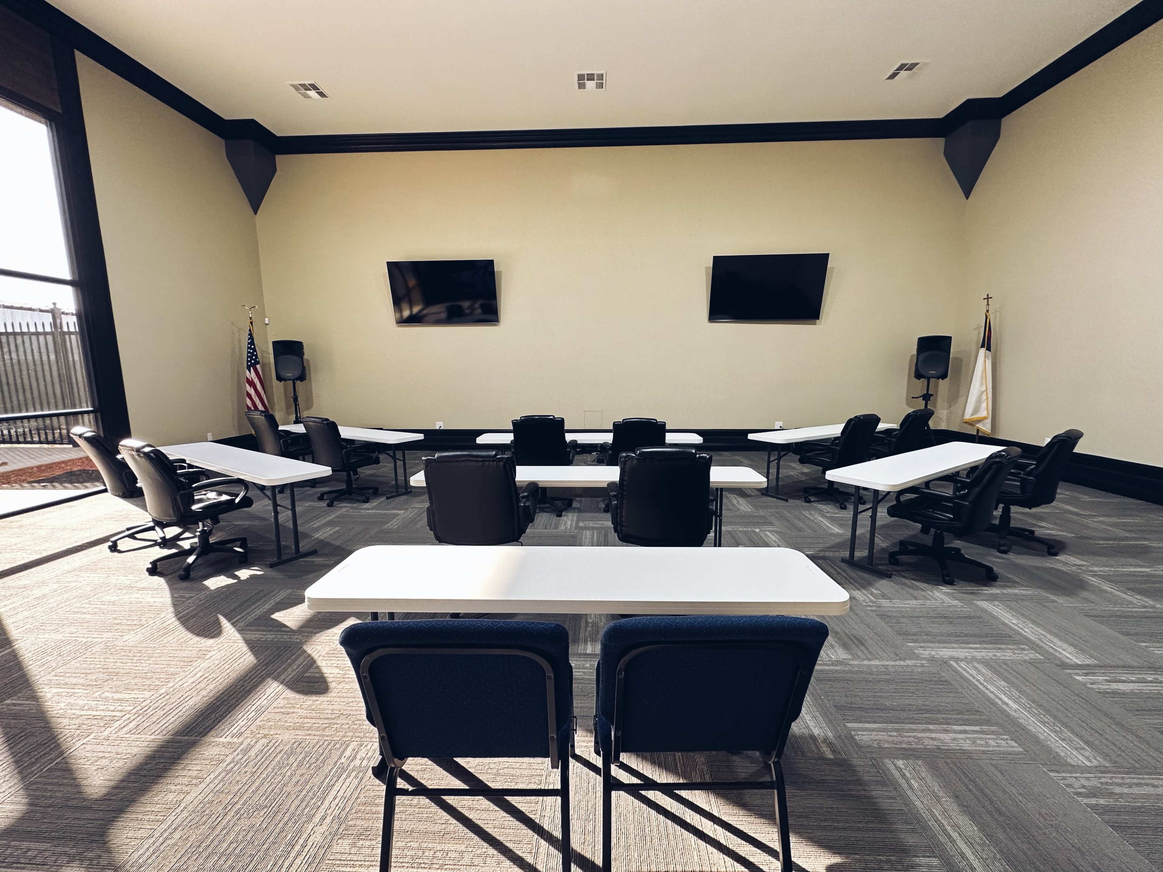 A conference room features several tables and chairs arranged in a U-shape, with two televisions mounted on the wall and windows allowing natural light.