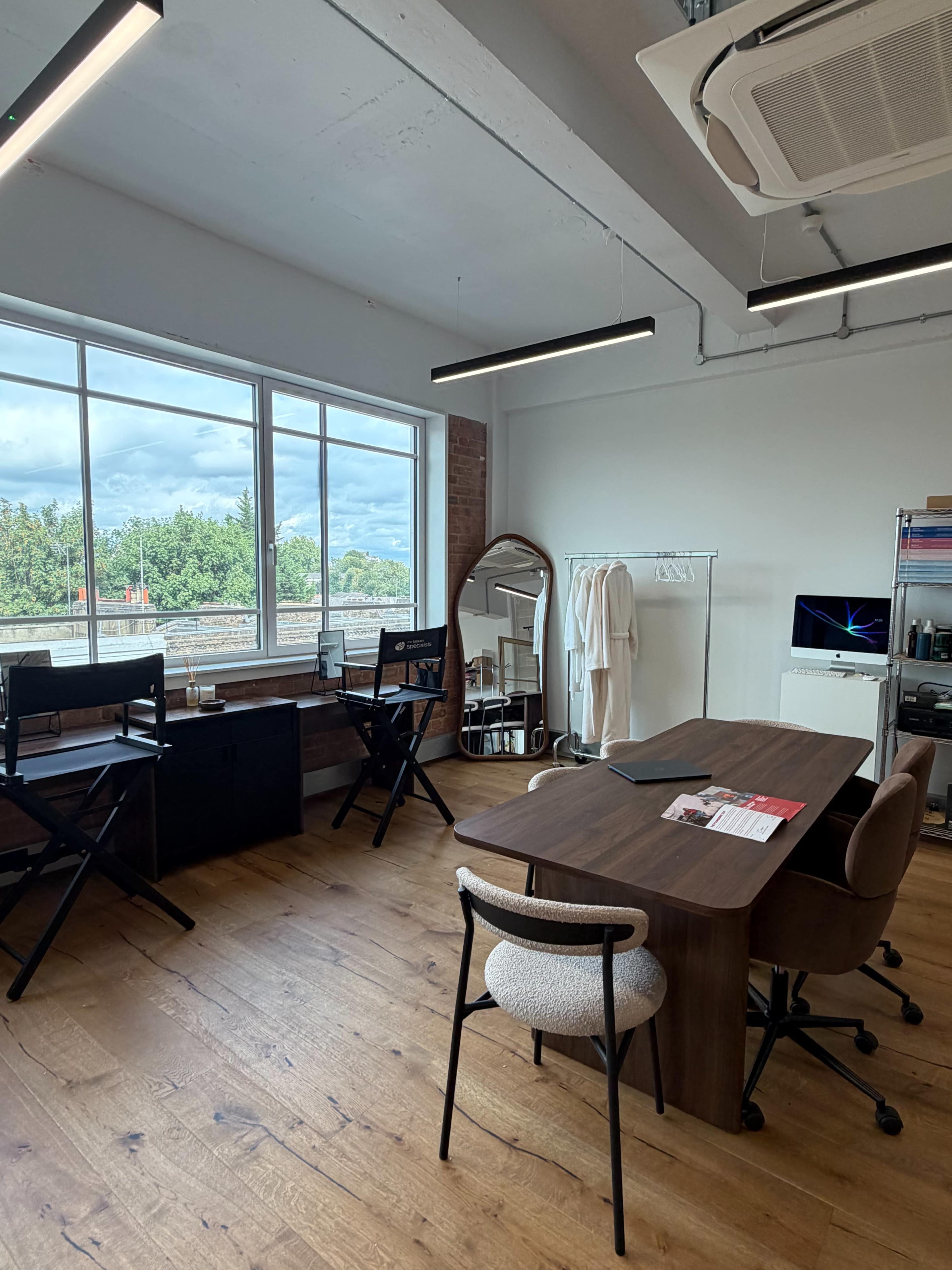 The image shows a modern office space featuring a wooden table, several chairs, a large window with a view of greenery outside, and minimalistic furniture.