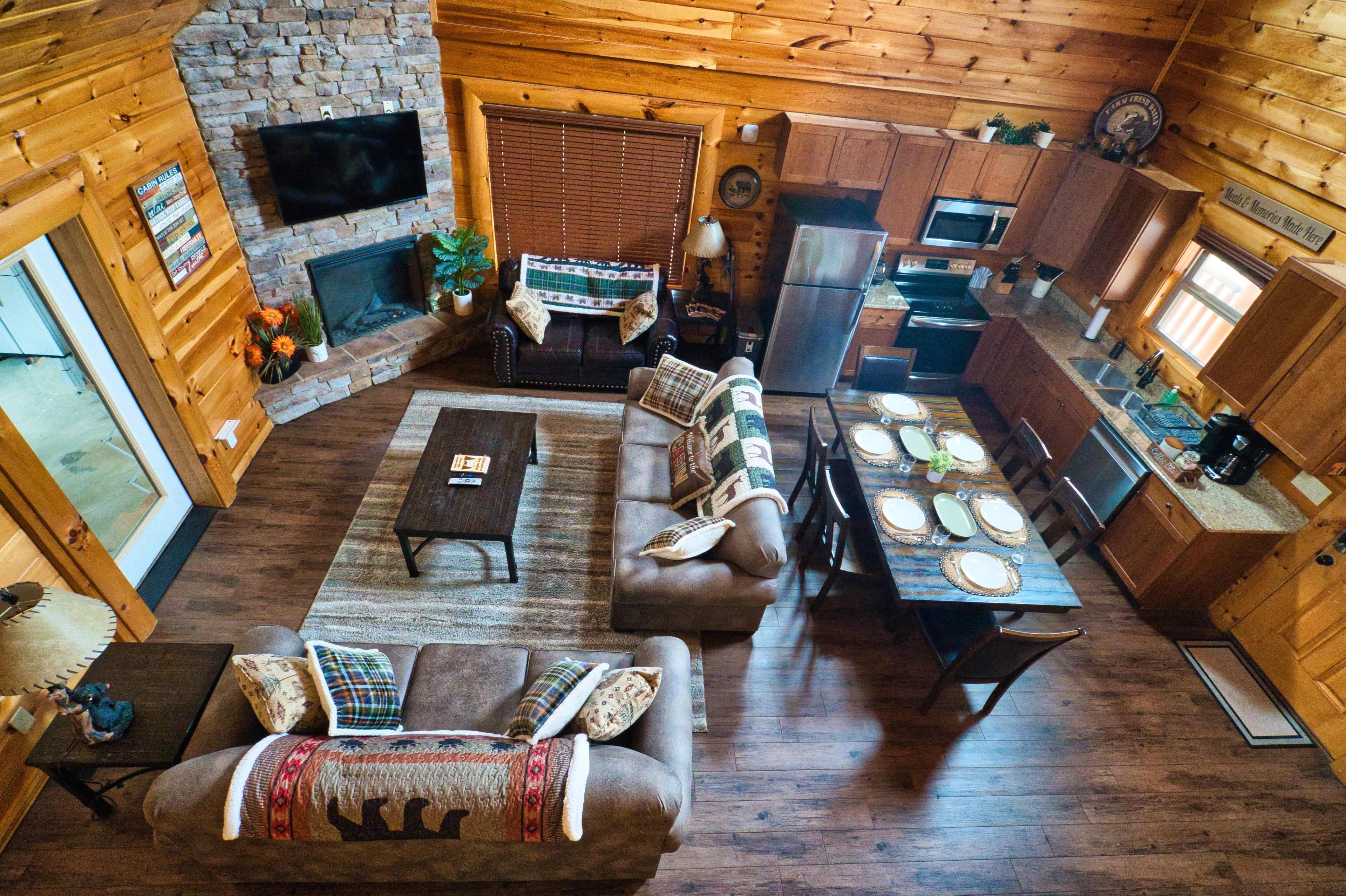 The image shows a spacious wooden cabin interior featuring a living area with two sofas, a coffee table, and a dining table, adjacent to a kitchen area and a stone fireplace.