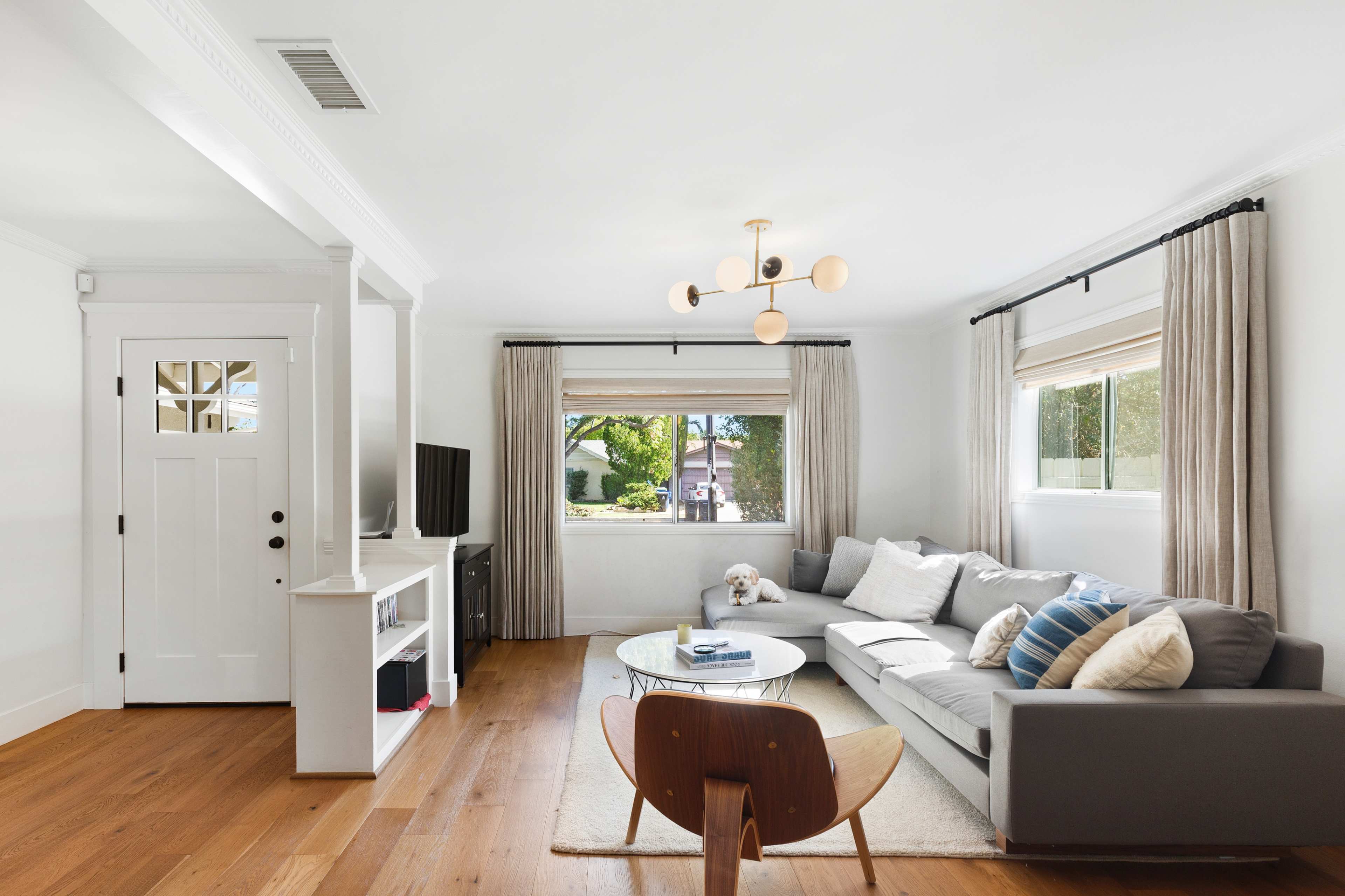 A modern living room features a gray sofa, a round glass coffee table, a television stand, and a light fixture, with large windows offering natural light and a view of greenery outside.