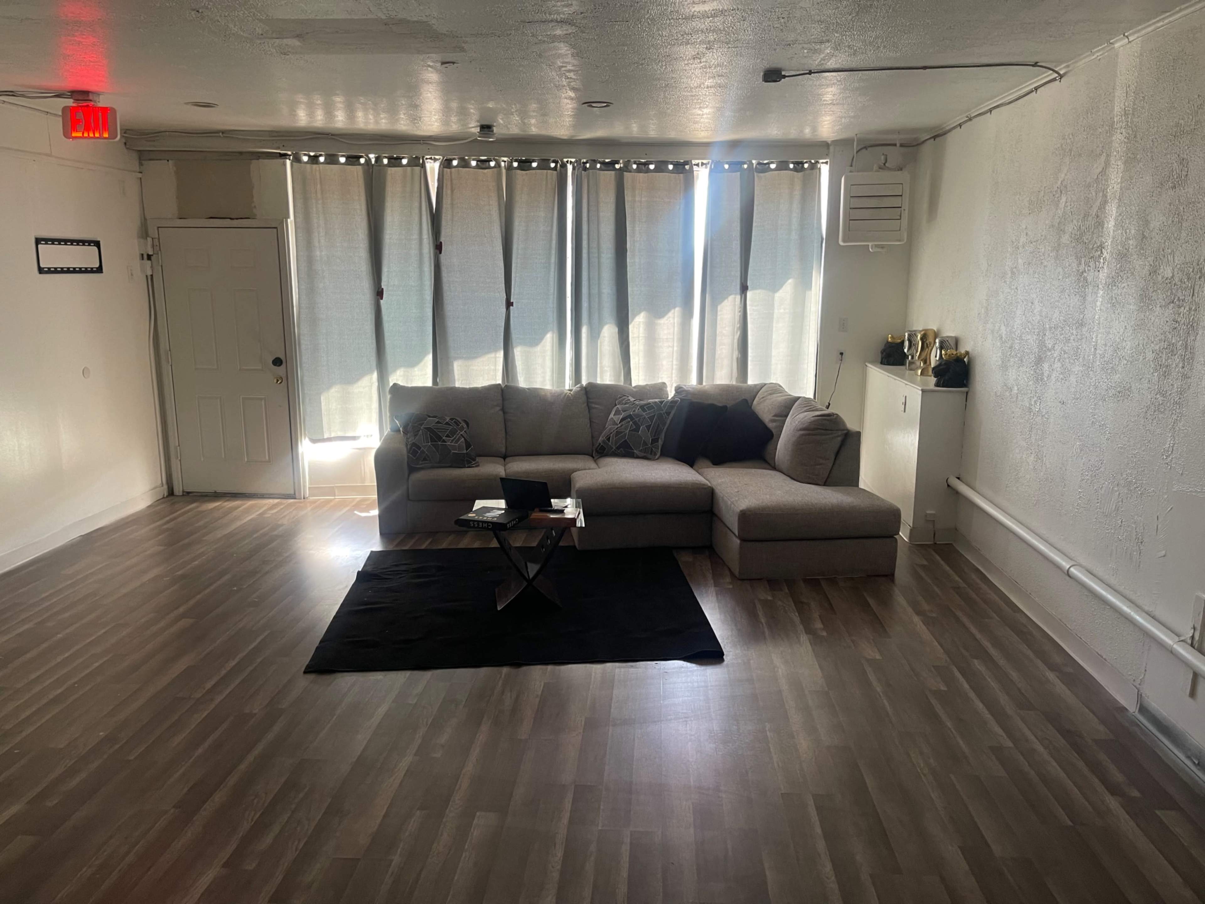 A spacious, minimally furnished living room featuring a large sectional sofa, a coffee table on a black rug, and natural light streaming through sheer curtains.