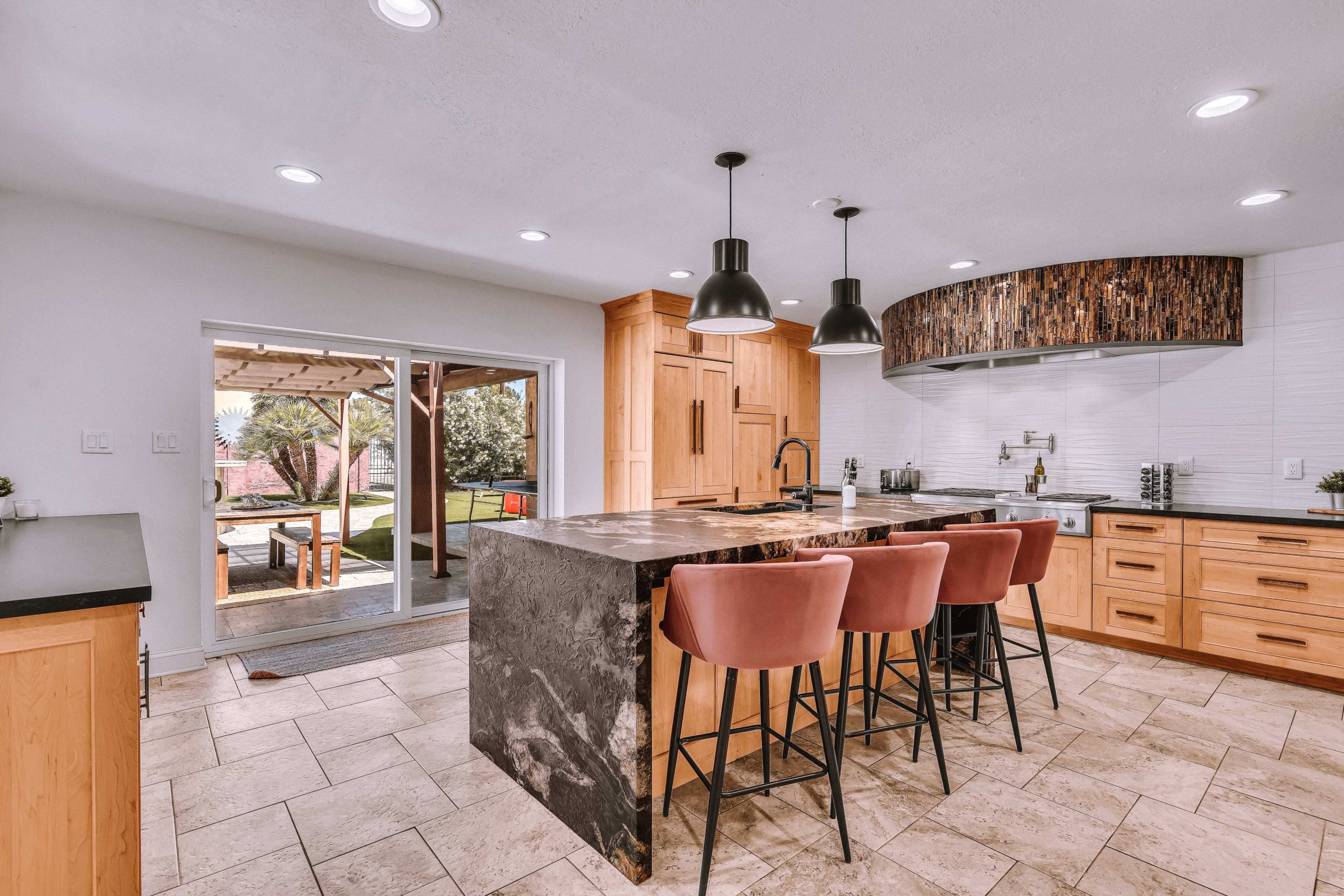 A modern kitchen featuring a large island with bar stools, light wood cabinetry, and a view of an outdoor patio through sliding glass doors.