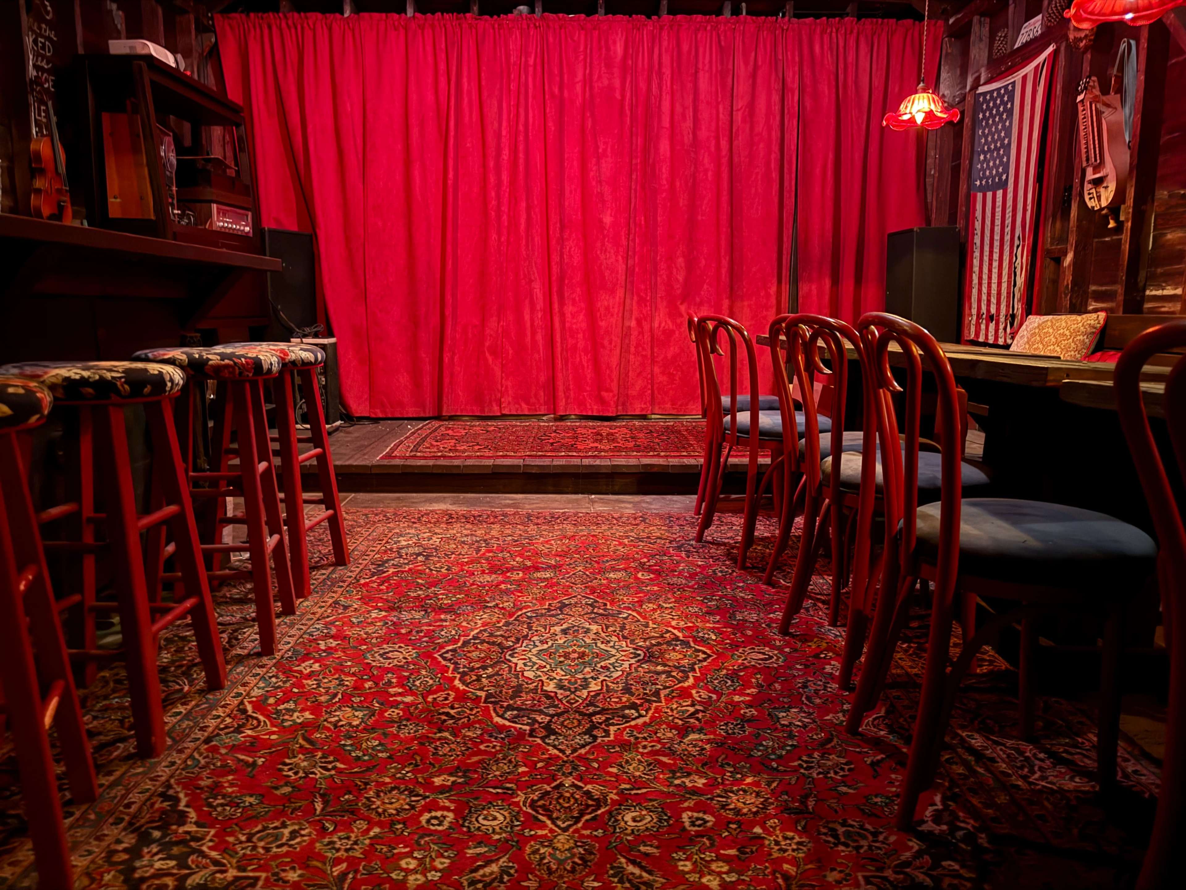 The image shows a cozy performance space with a red curtain, wooden bar stools, and patterned carpeting.