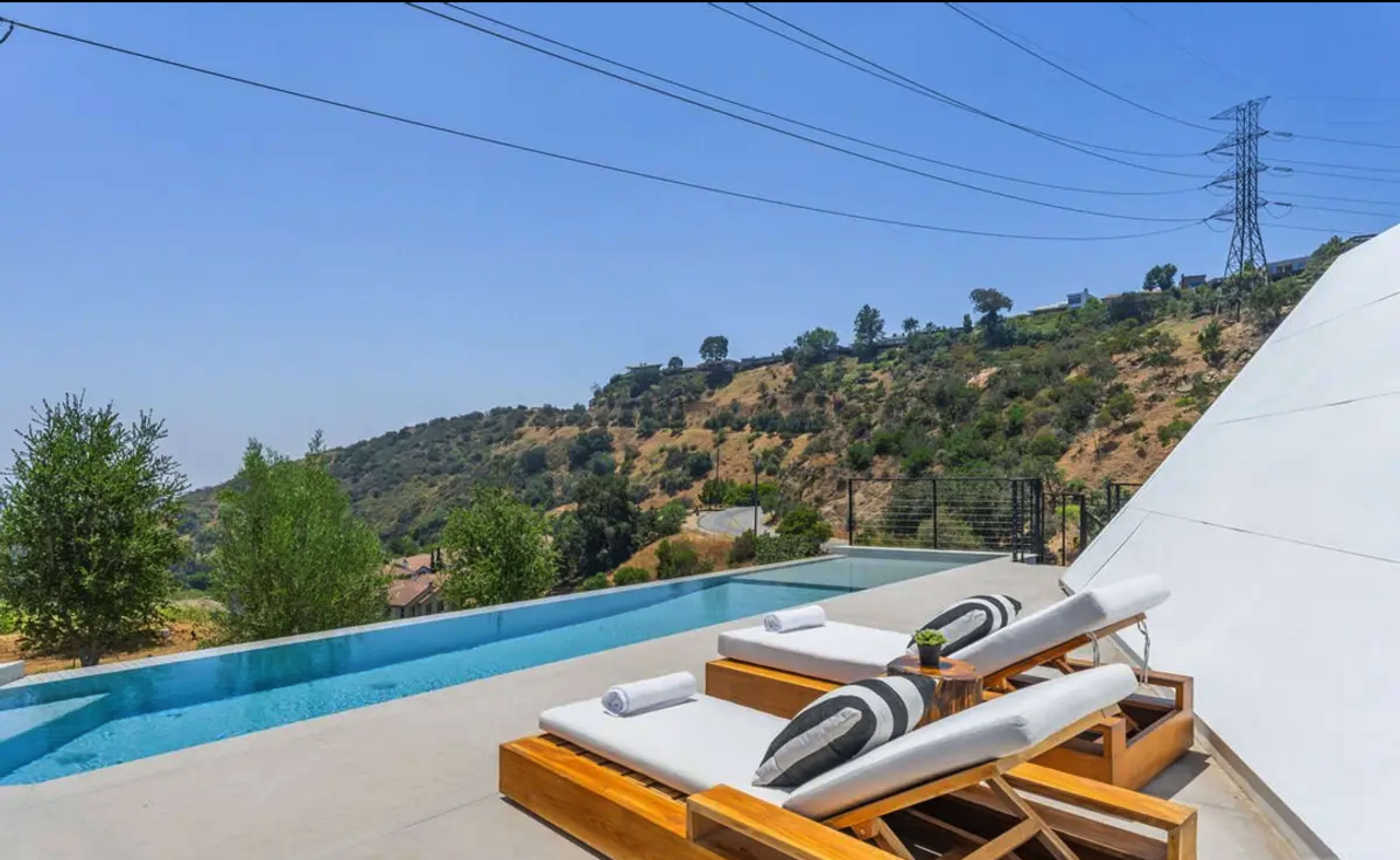 A pair of lounge chairs are positioned beside a swimming pool that overlooks a hillside landscape under a clear blue sky.