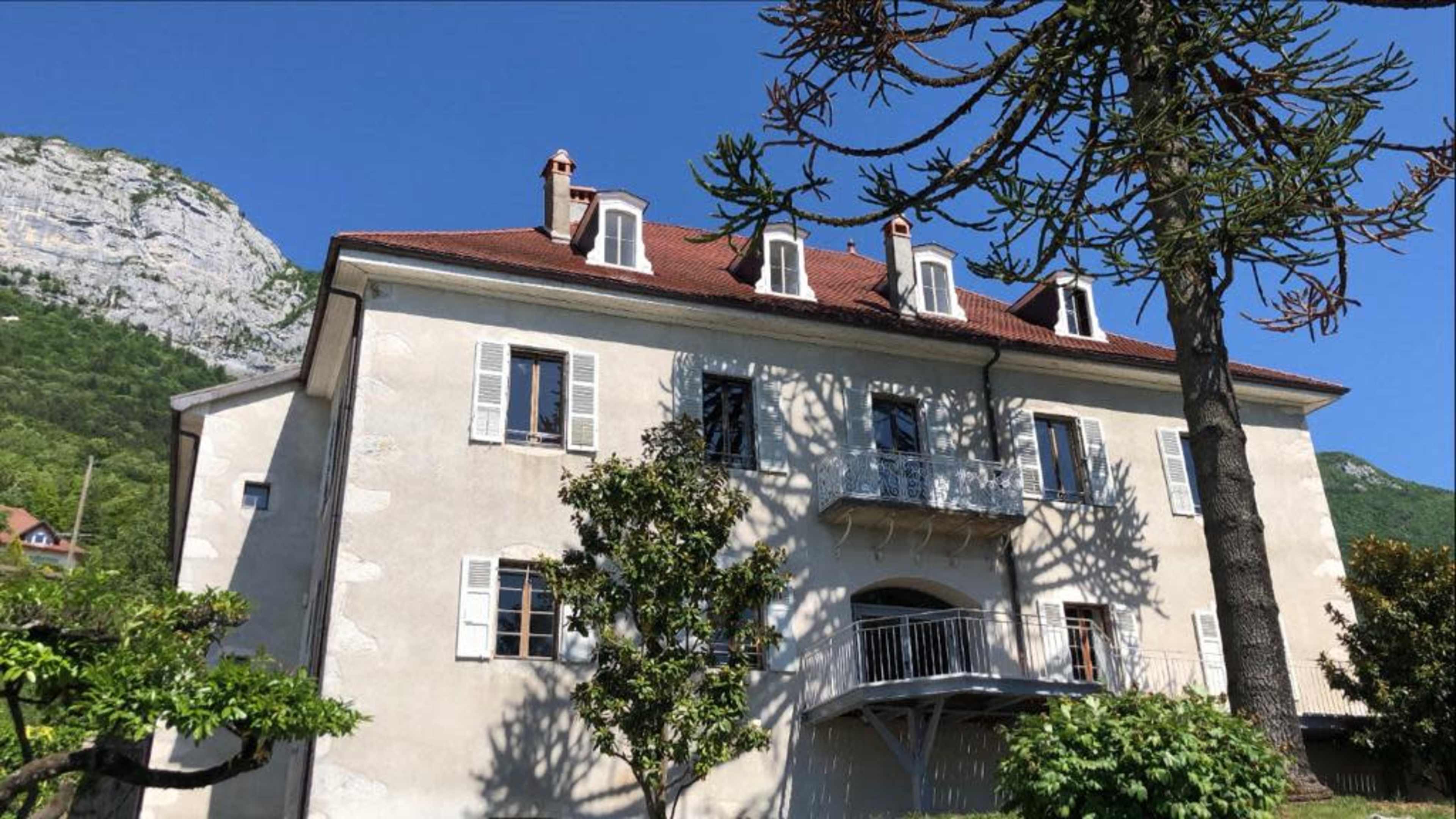A three-story house with a balcony and shutters is situated against a backdrop of mountains and greenery.