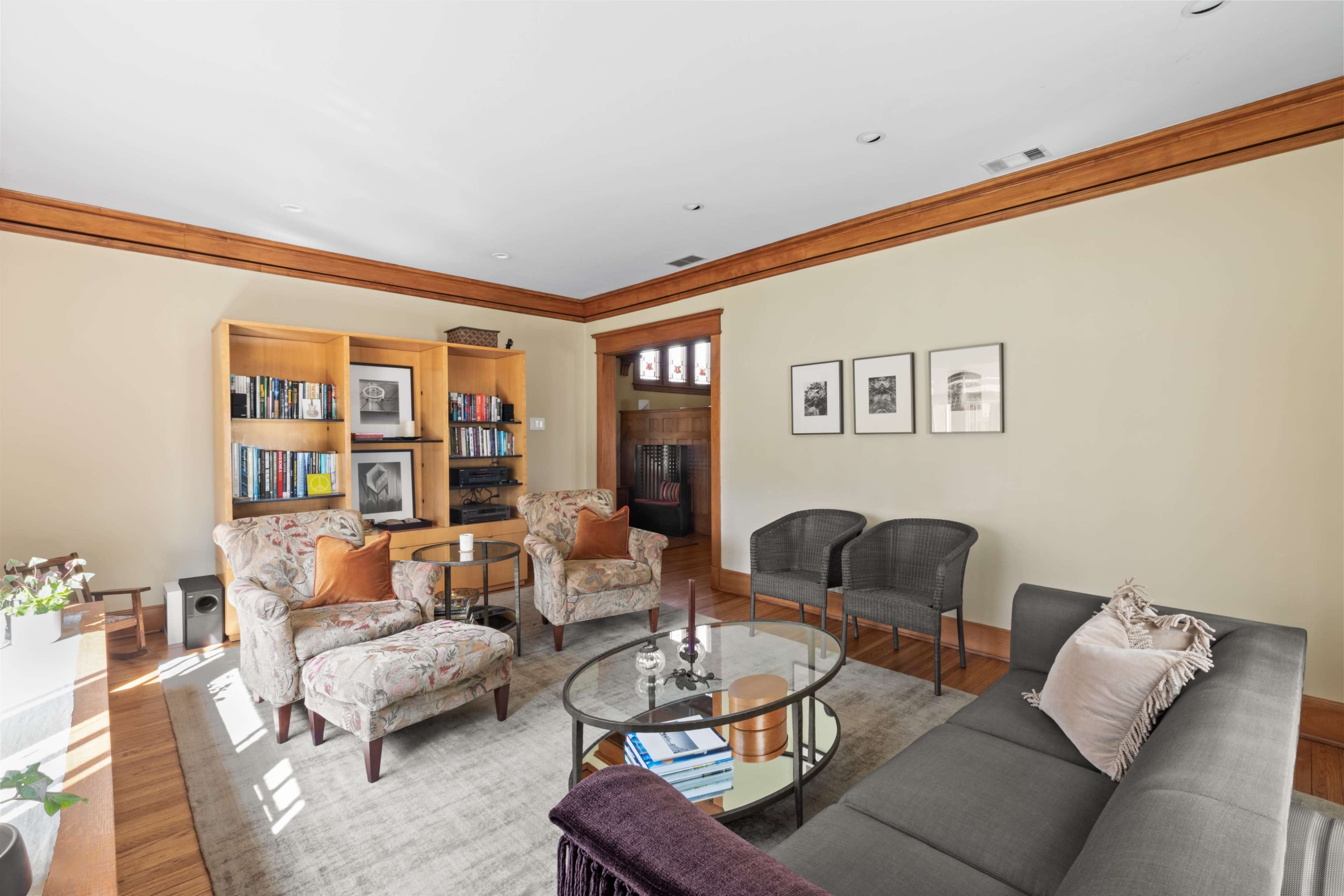 The image shows a living room featuring two floral armchairs, a gray sofa, a glass coffee table, and a bookshelf filled with books against a beige wall.