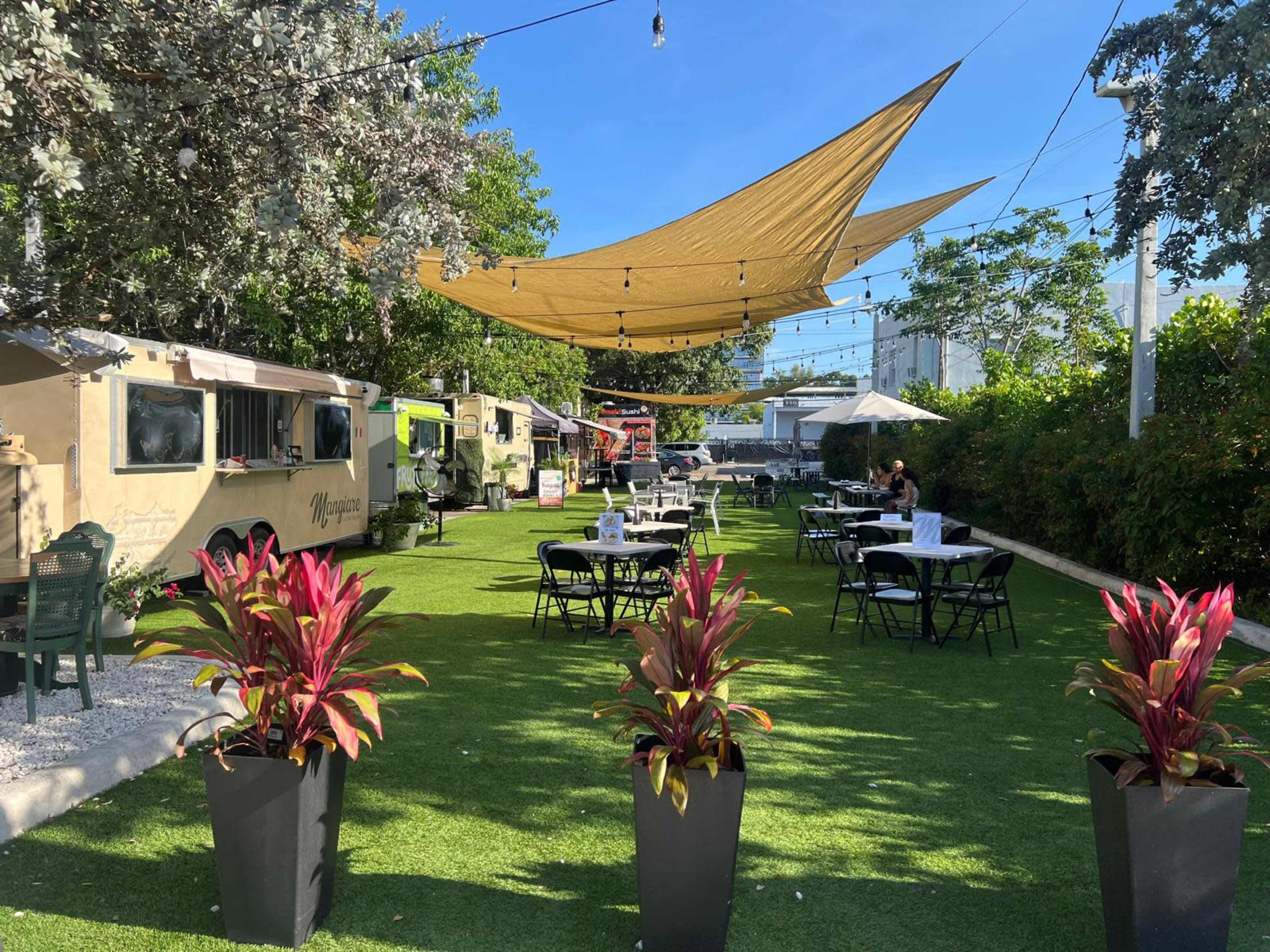 The image shows an outdoor dining area featuring food trucks, tables with umbrellas, and decorative plants under a sunny blue sky.