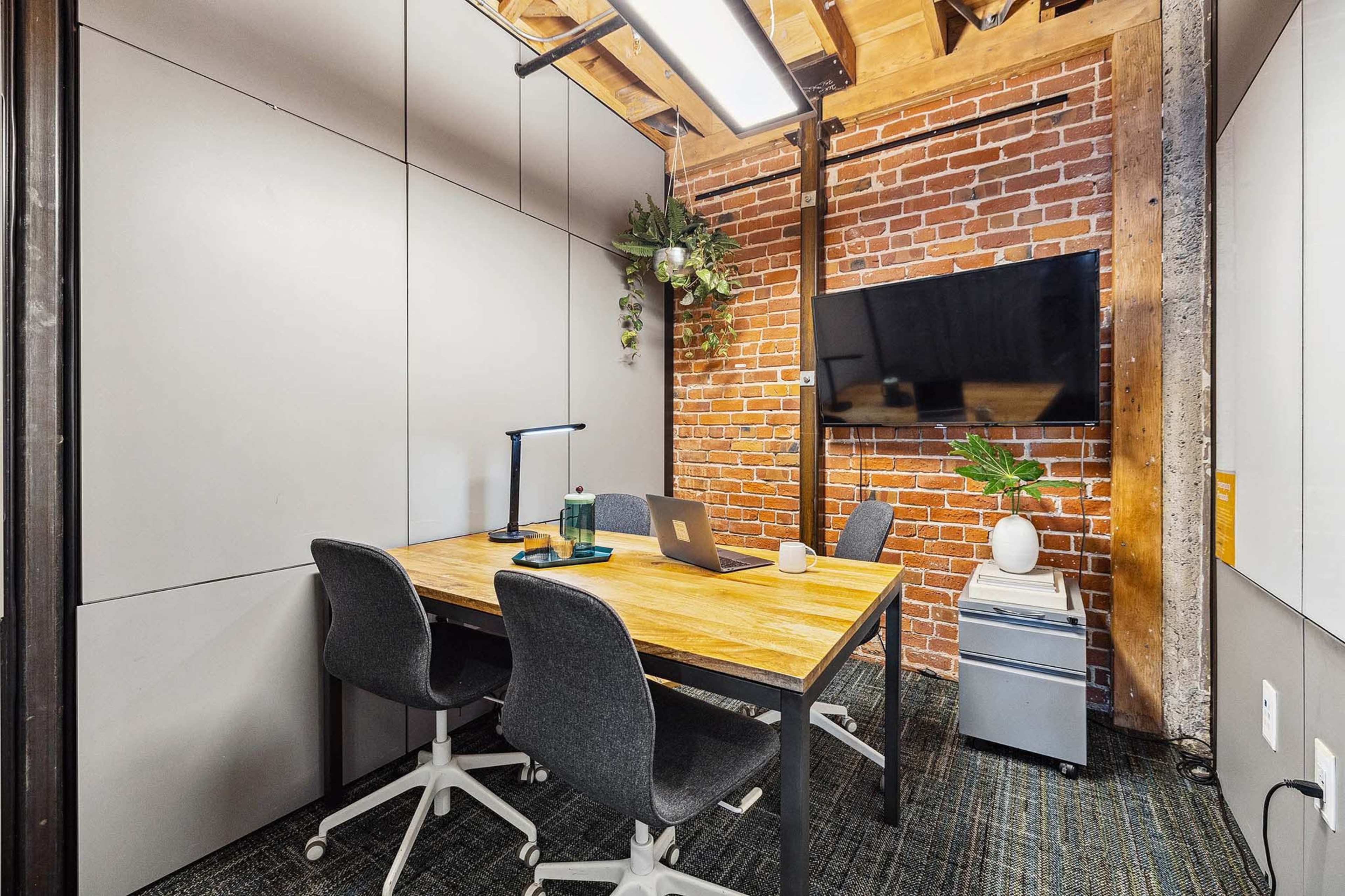 A small meeting room features a wooden table, three chairs, a large wall-mounted TV, and exposed brick walls with greenery decor.
