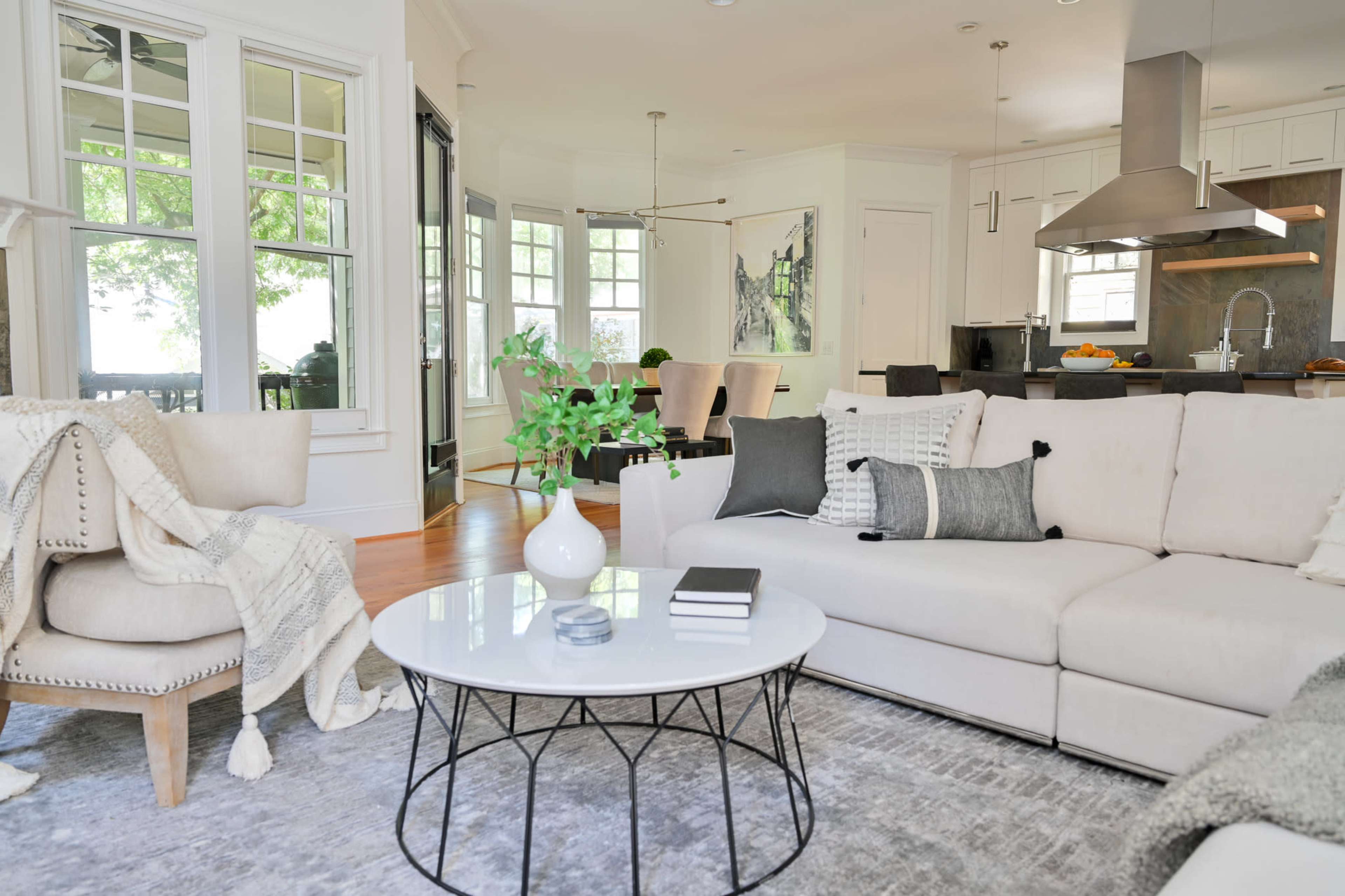 A light-filled living room with a white sectional sofa, a round glass coffee table, and large windows overlooking greenery.