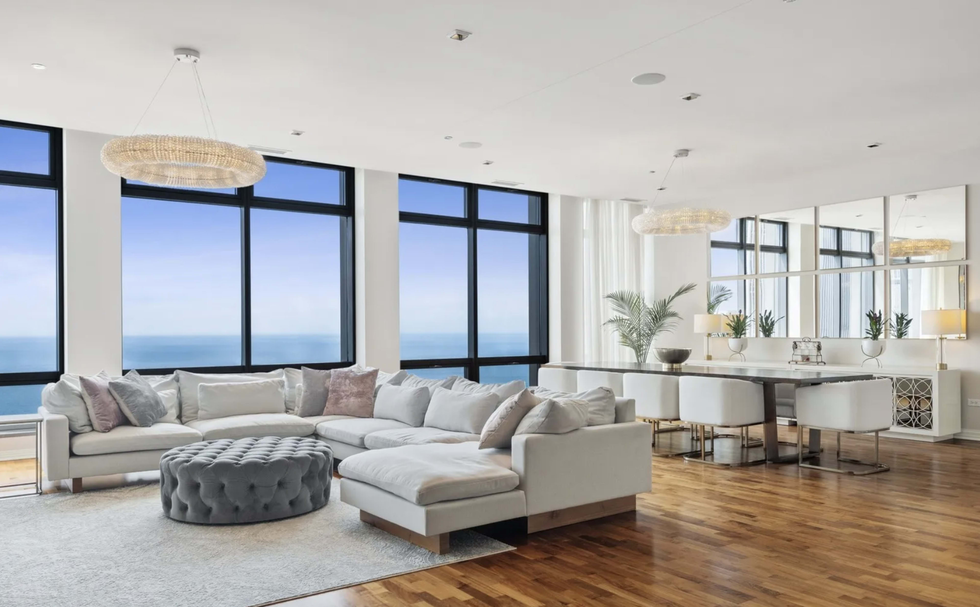 The image shows a modern living room with large windows offering an ocean view, featuring a sectional sofa, a round ottoman, and a dining area with white chairs.