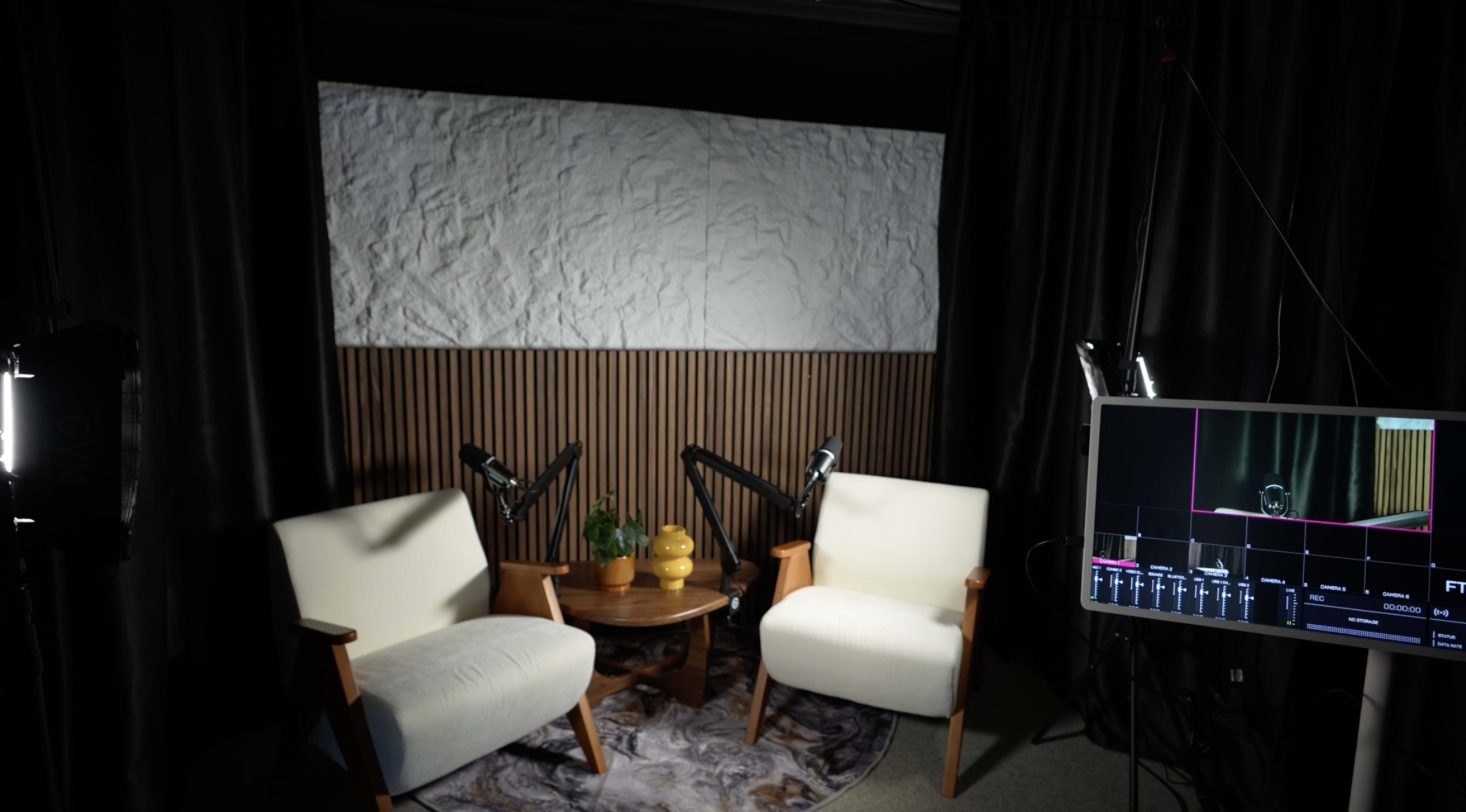 The image shows a well-lit indoor setting with two white chairs positioned across from a small round table, surrounded by acoustic treatment and a blank screen in the background.
