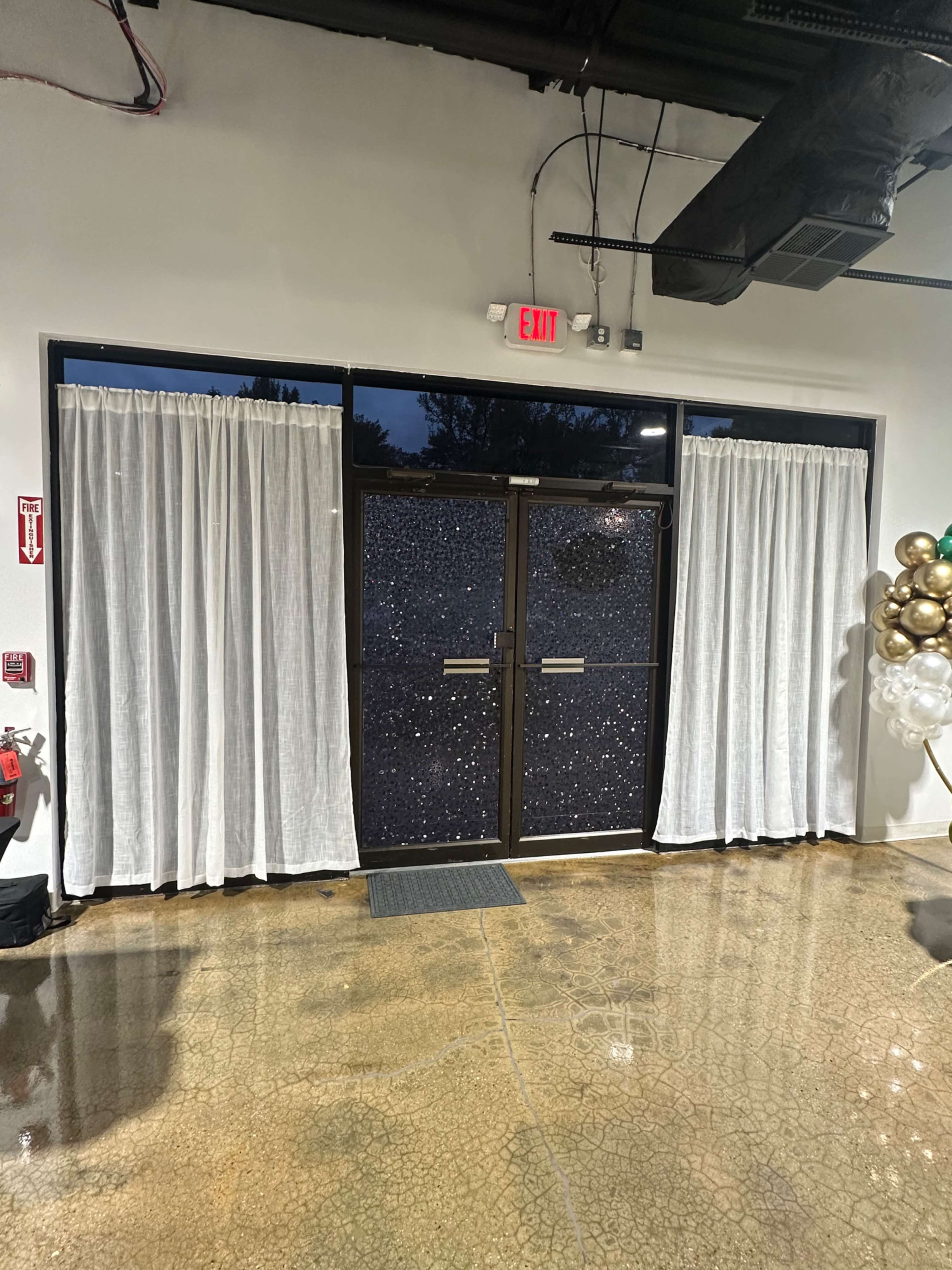 The image shows a set of double glass doors with a textured surface, flanked by white curtains, and an exit sign above in a well-lit indoor space with polished concrete flooring.