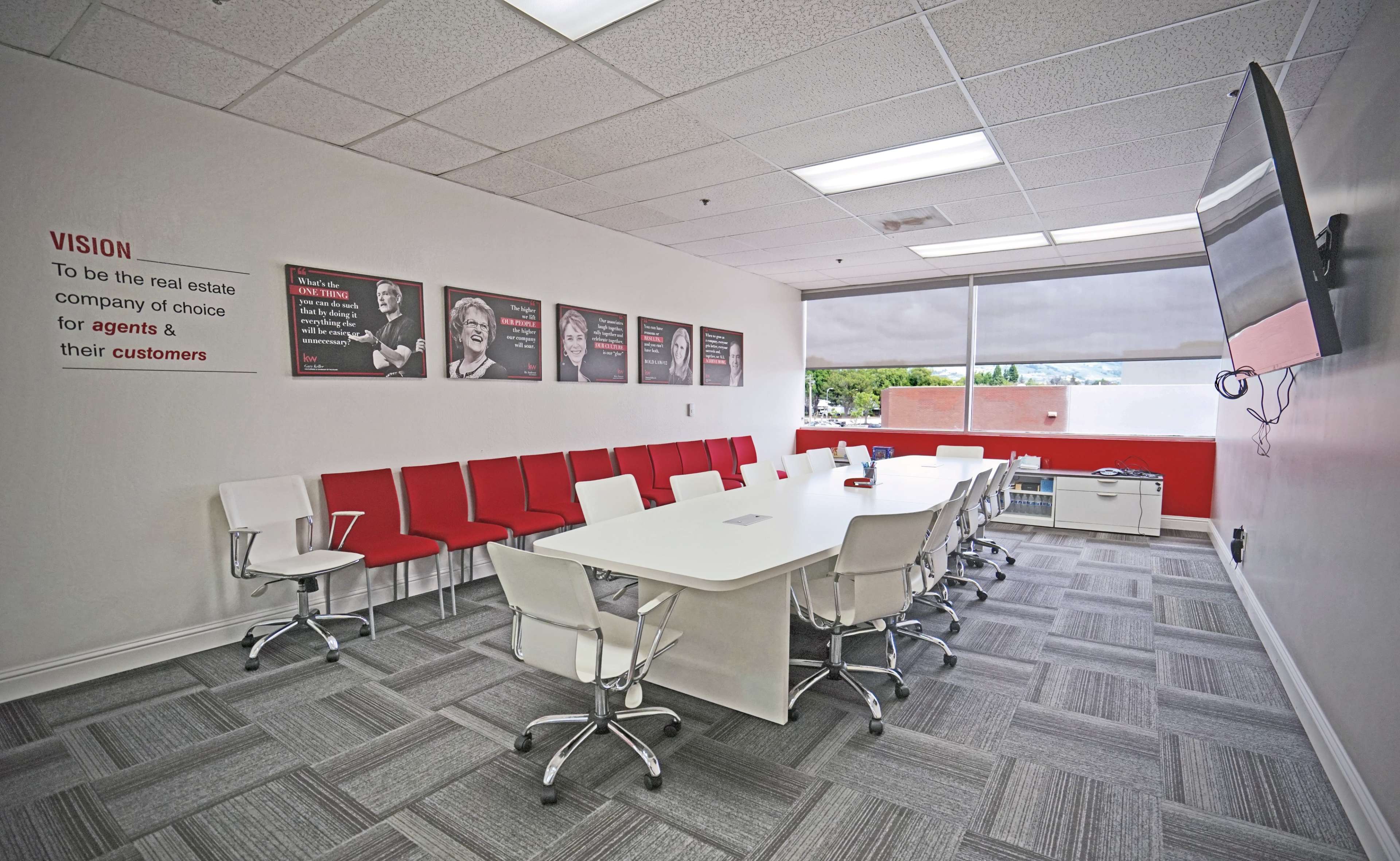 A modern conference room features a large white table surrounded by ergonomic chairs, with a wall displaying framed portraits and a vision statement.