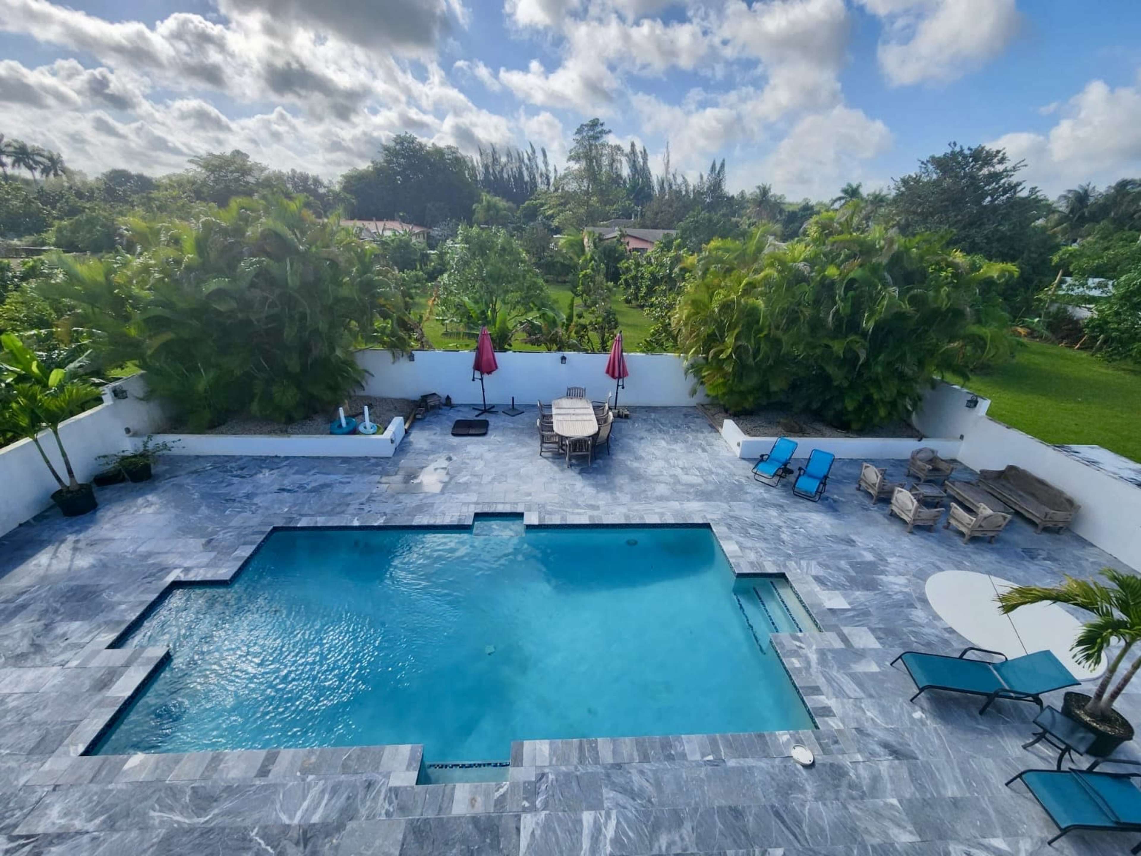 A large rectangular swimming pool surrounded by lounge chairs and tables, set in a landscaped backyard with lush greenery and palm trees.