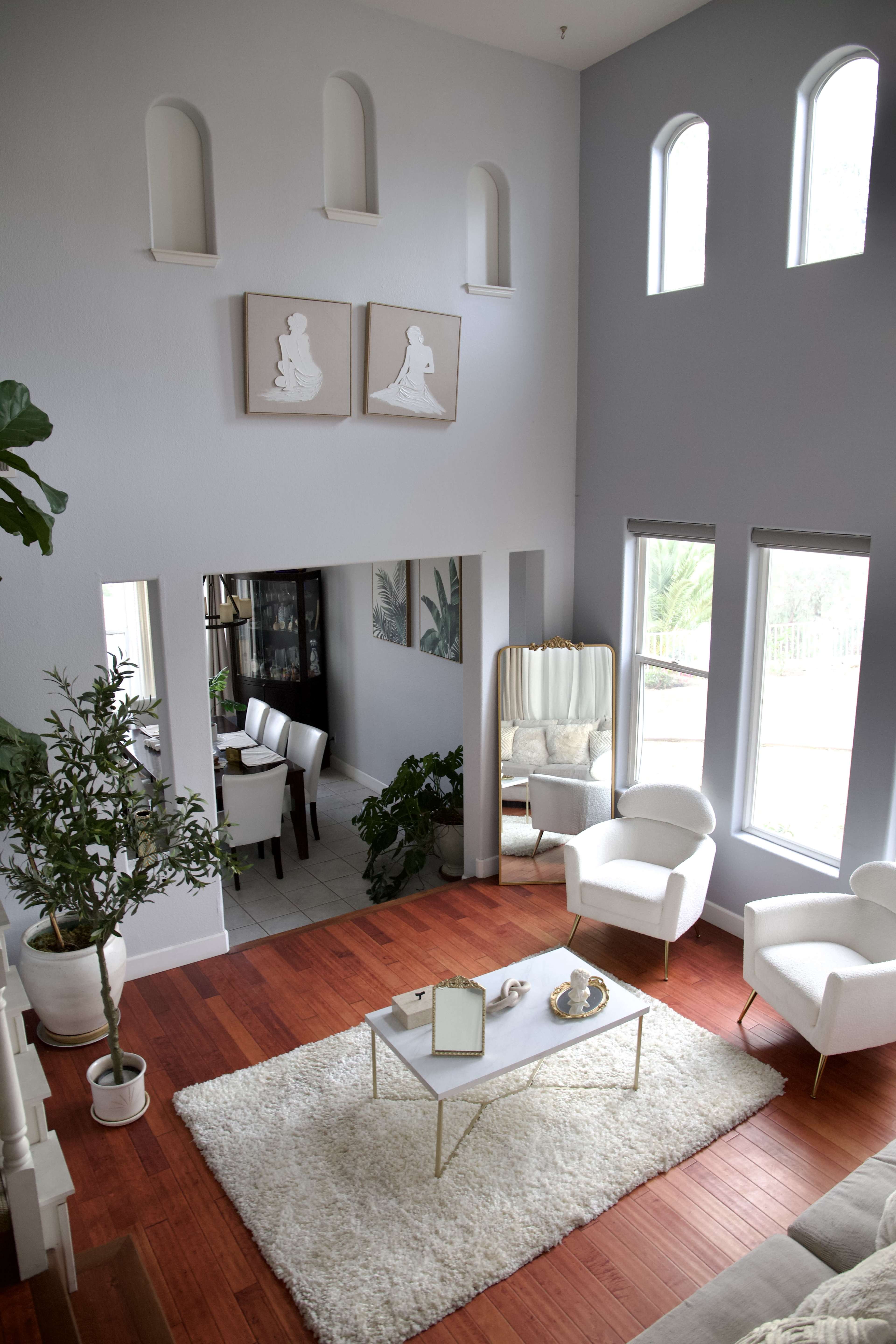 The image shows a spacious living room with a modern decor featuring a large white rug, a coffee table, and two white chairs, with large windows allowing natural light to fill the space.
