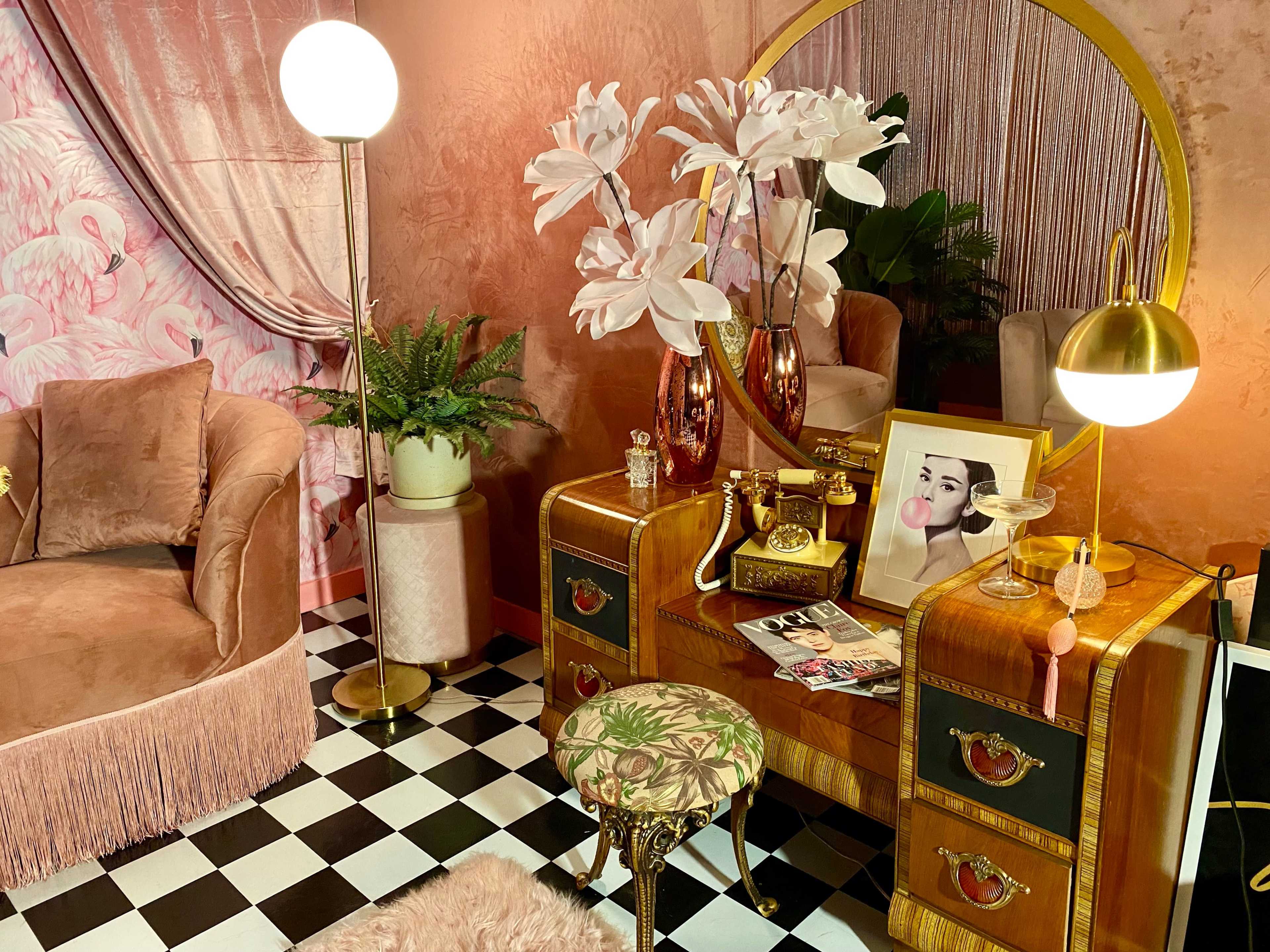 The scene features a stylish interior with a vintage dresser, decorative flowers in vases, a green plant, a round mirror, and a plush couch against a pink backdrop.