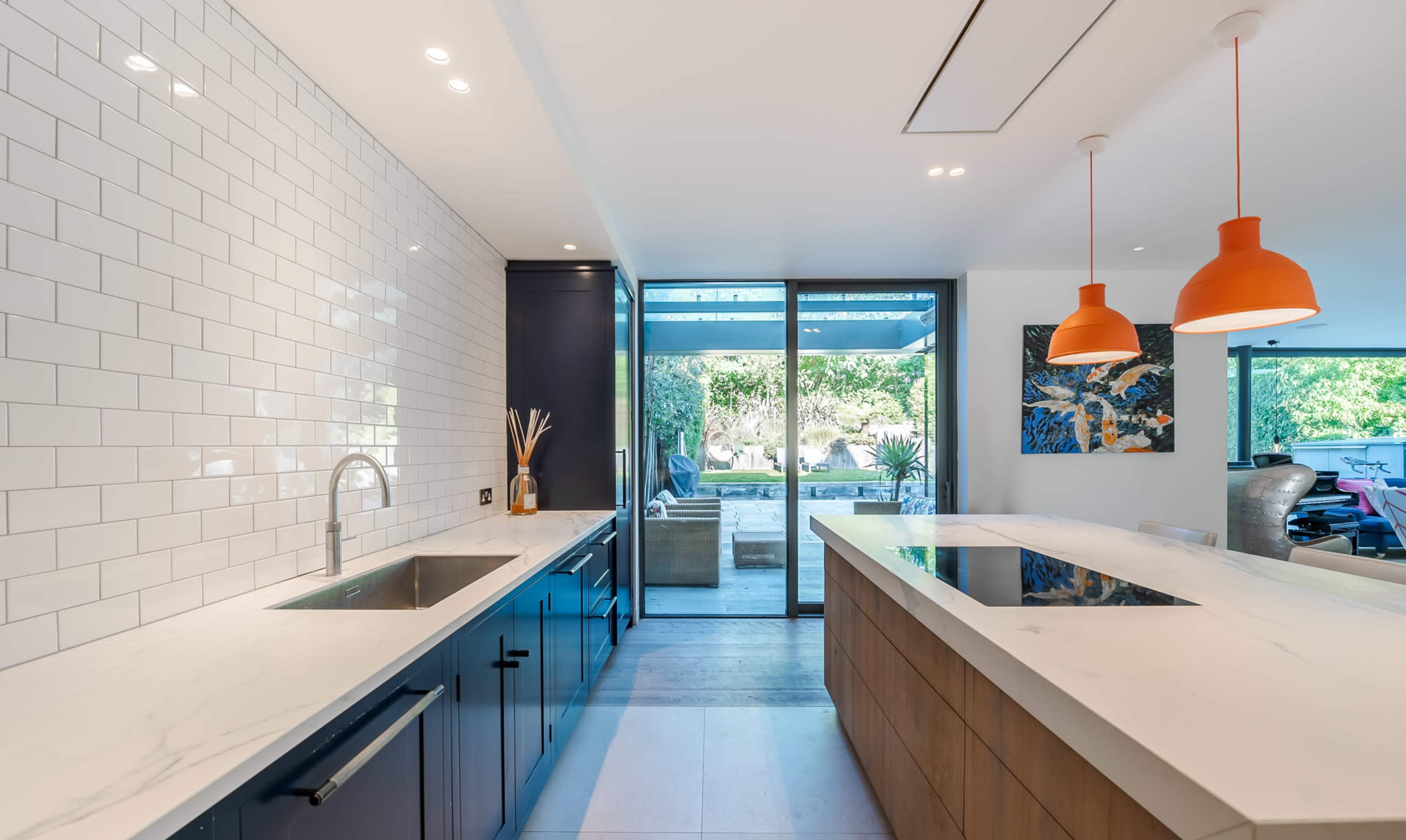 The kitchen features a large island with a mix of light and dark cabinetry, a stainless steel sink, and orange pendant lights, overlooking an outdoor area through glass sliding doors.