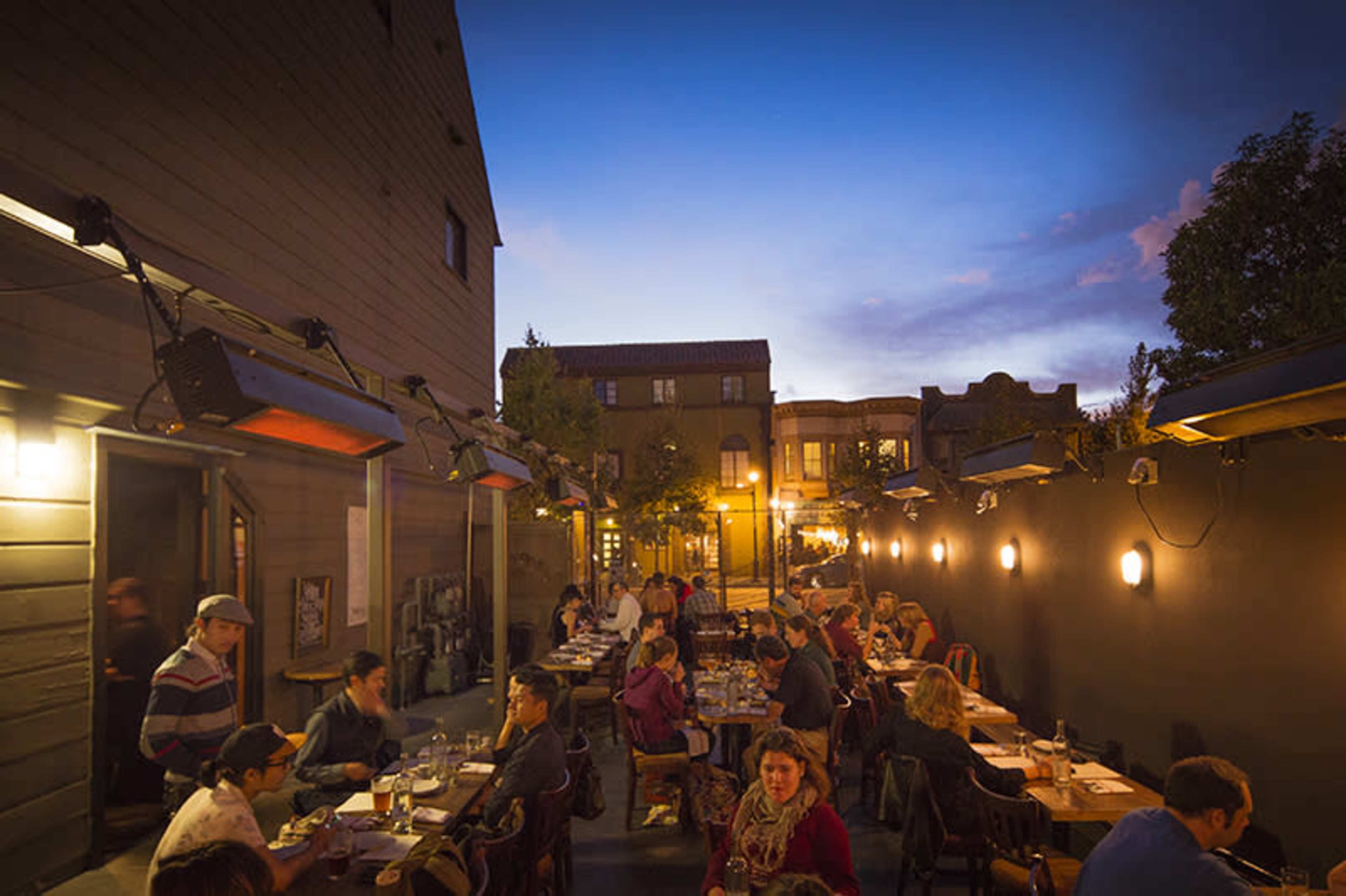 A bustling outdoor restaurant scene is captured at dusk, featuring diners seated at long tables under warm lighting.