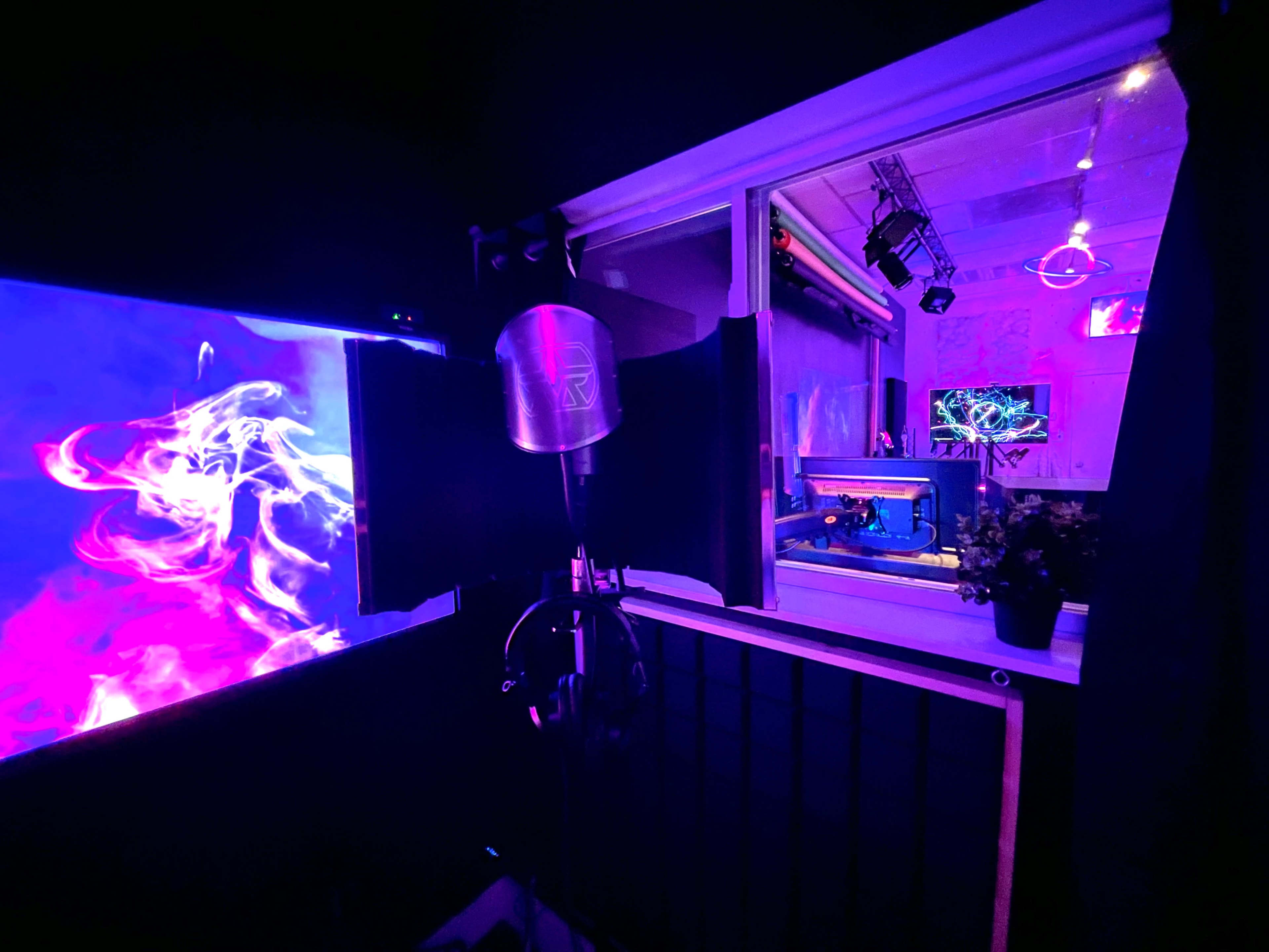 A recording studio with a microphone in front of a colorful visual display and a window revealing more equipment and lighting inside.
