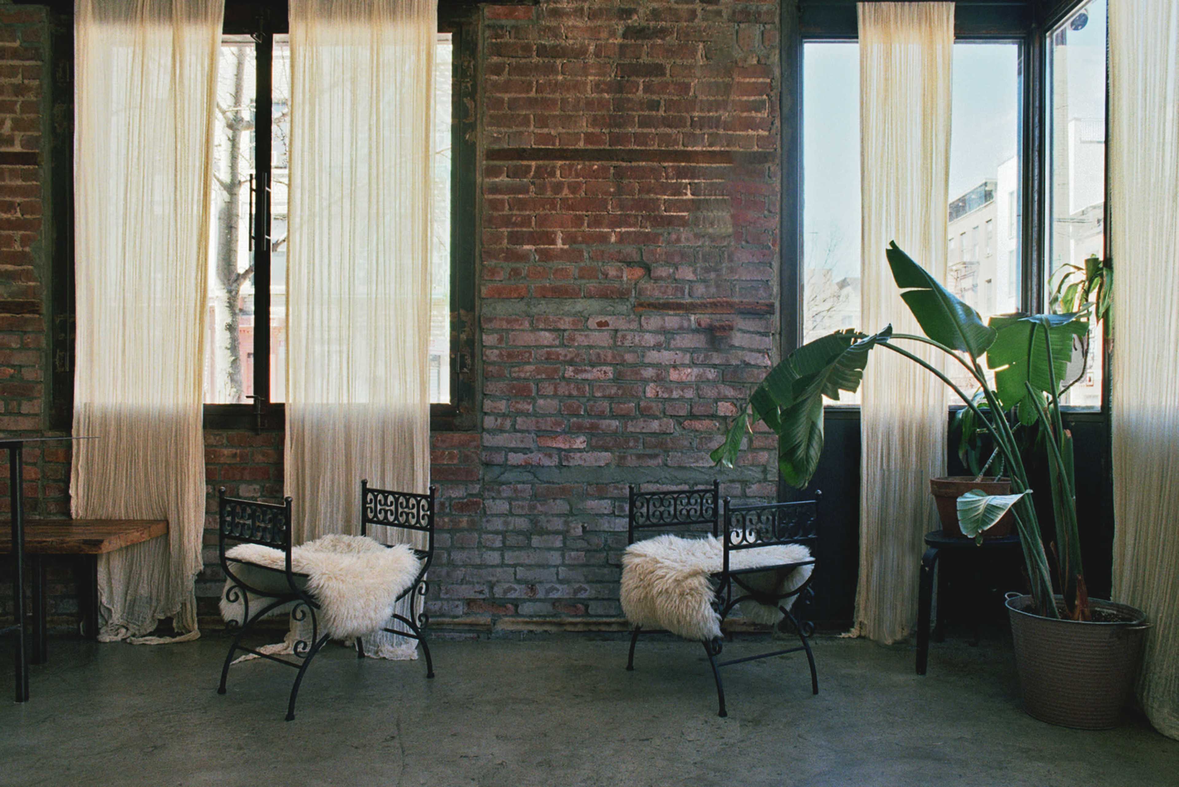 The image features two iron chairs with fur cushions positioned beside large windows adorned with sheer curtains, set against a backdrop of exposed brick walls and a potted plant.