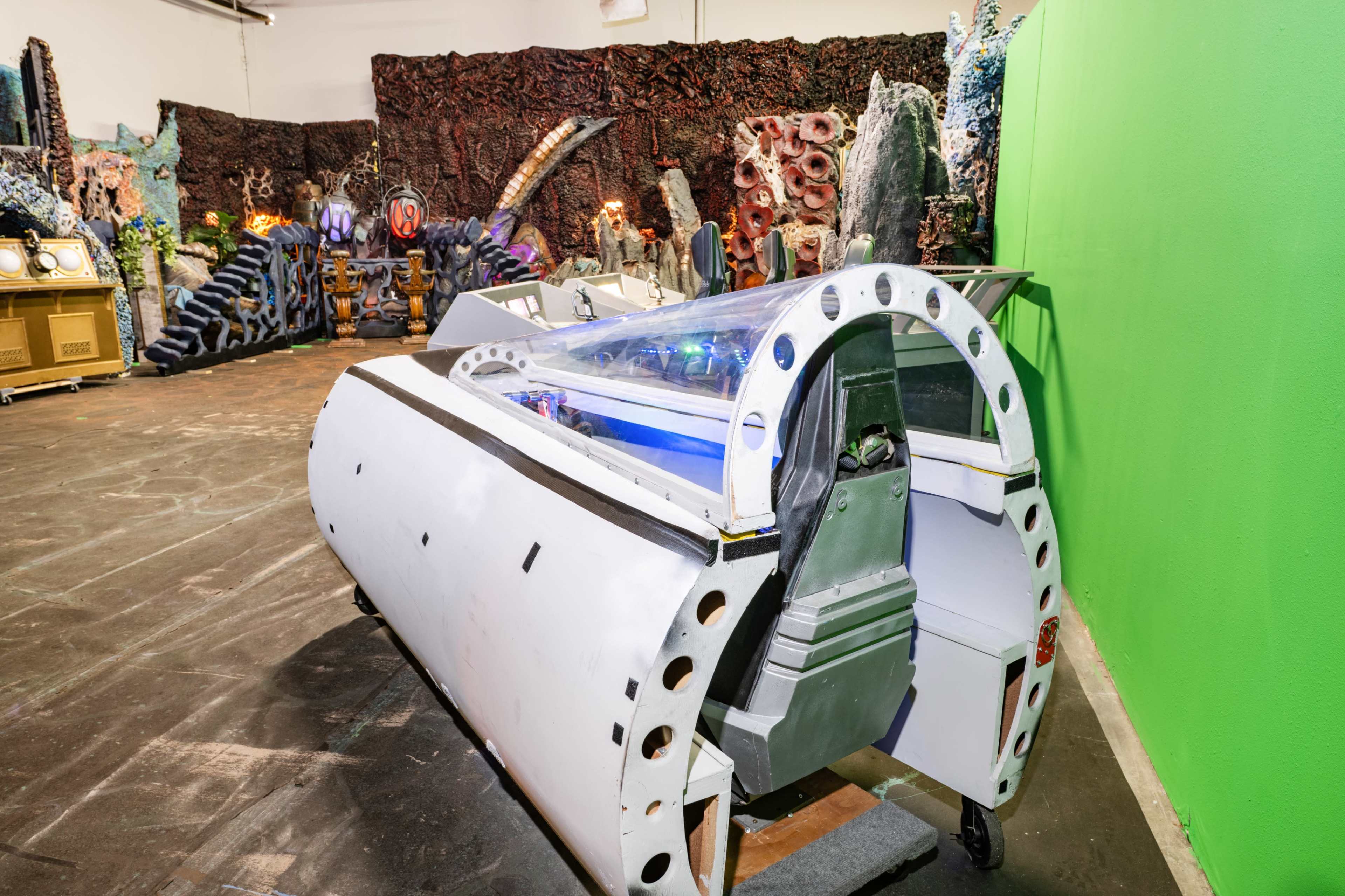 The image shows a futuristic machine with a transparent dome, set against a backdrop of textured, colorful walls and sci-fi-themed decorations.