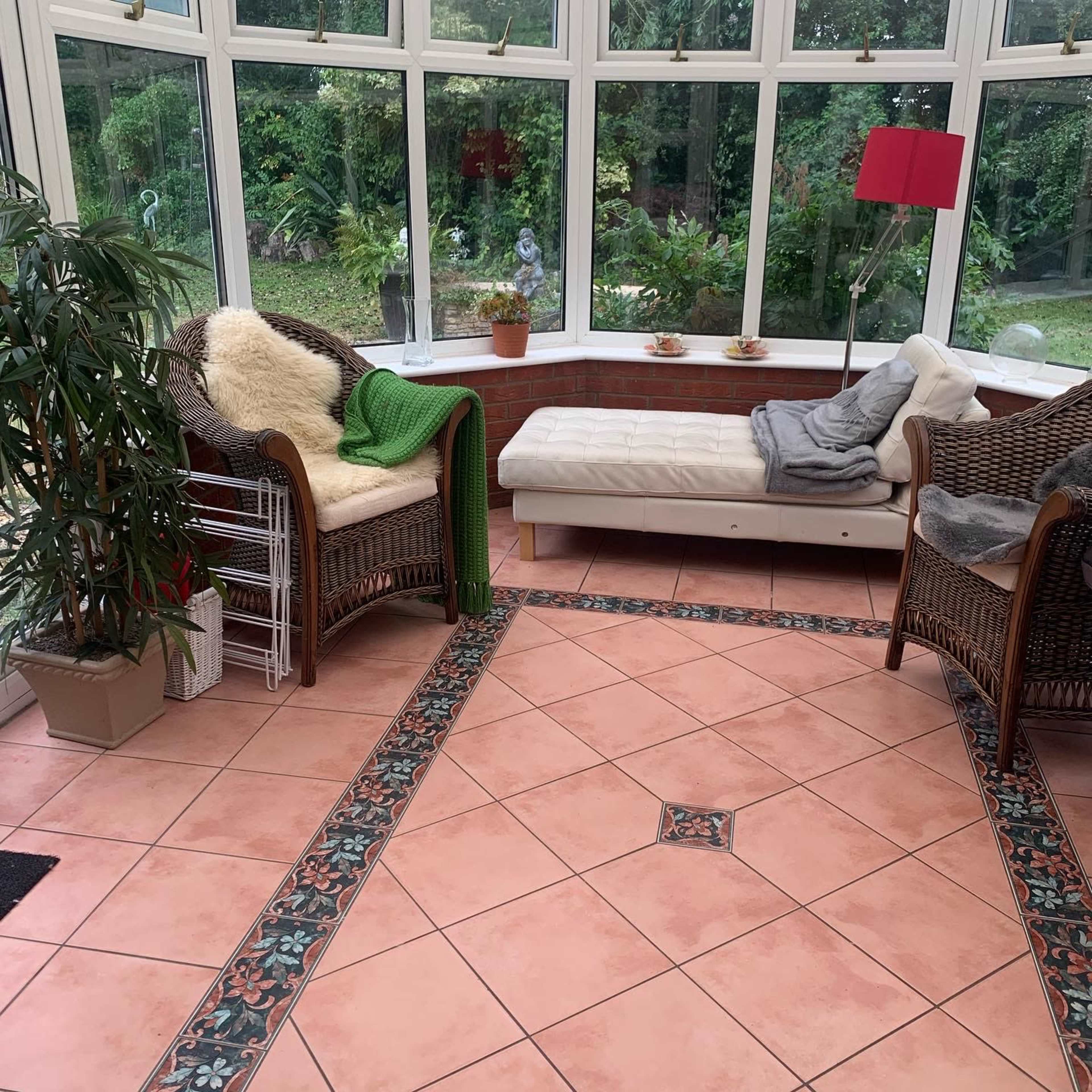 The image shows a bright sunroom with large windows, a tiled floor featuring a floral border, two wicker chairs, and a white daybed with cushions.