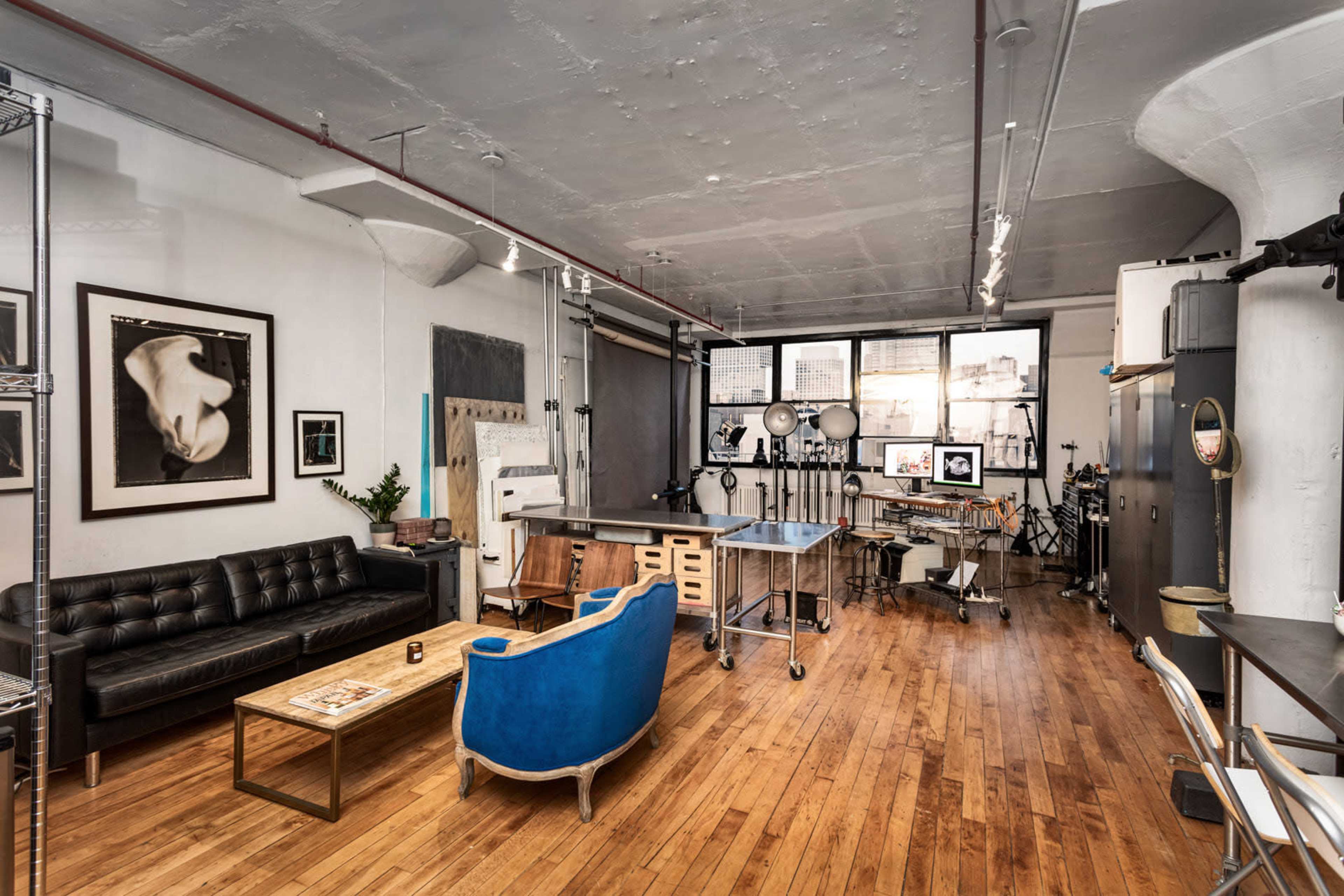 The image depicts a spacious studio with wooden floors, featuring a black leather sofa, a blue upholstered chair, and various art equipment along the walls.