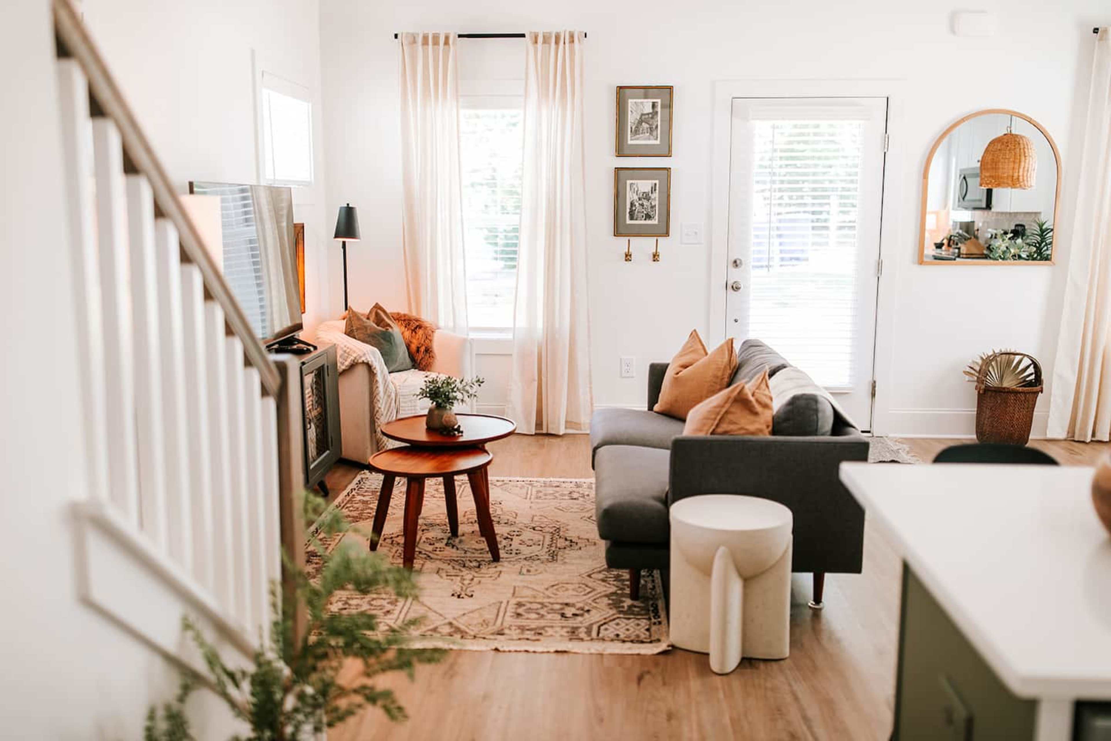 The scene features a bright living room with a gray sofa, an armchair, a round coffee table, and a television against a white wall, complemented by light curtains and decorative plants.