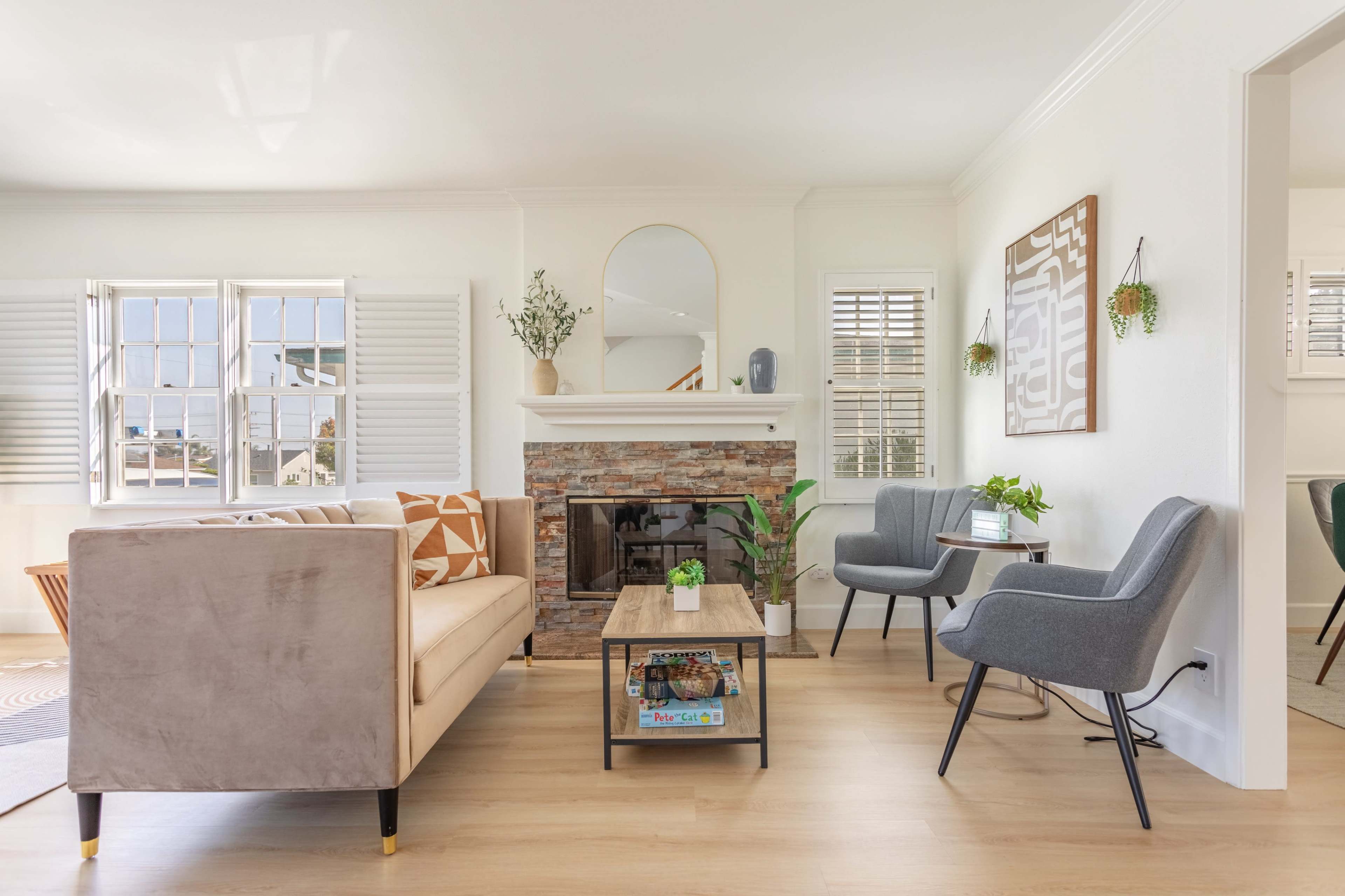 The image shows a modern living room featuring a beige sofa, a stone fireplace, and two gray chairs, with natural light streaming through the windows.
