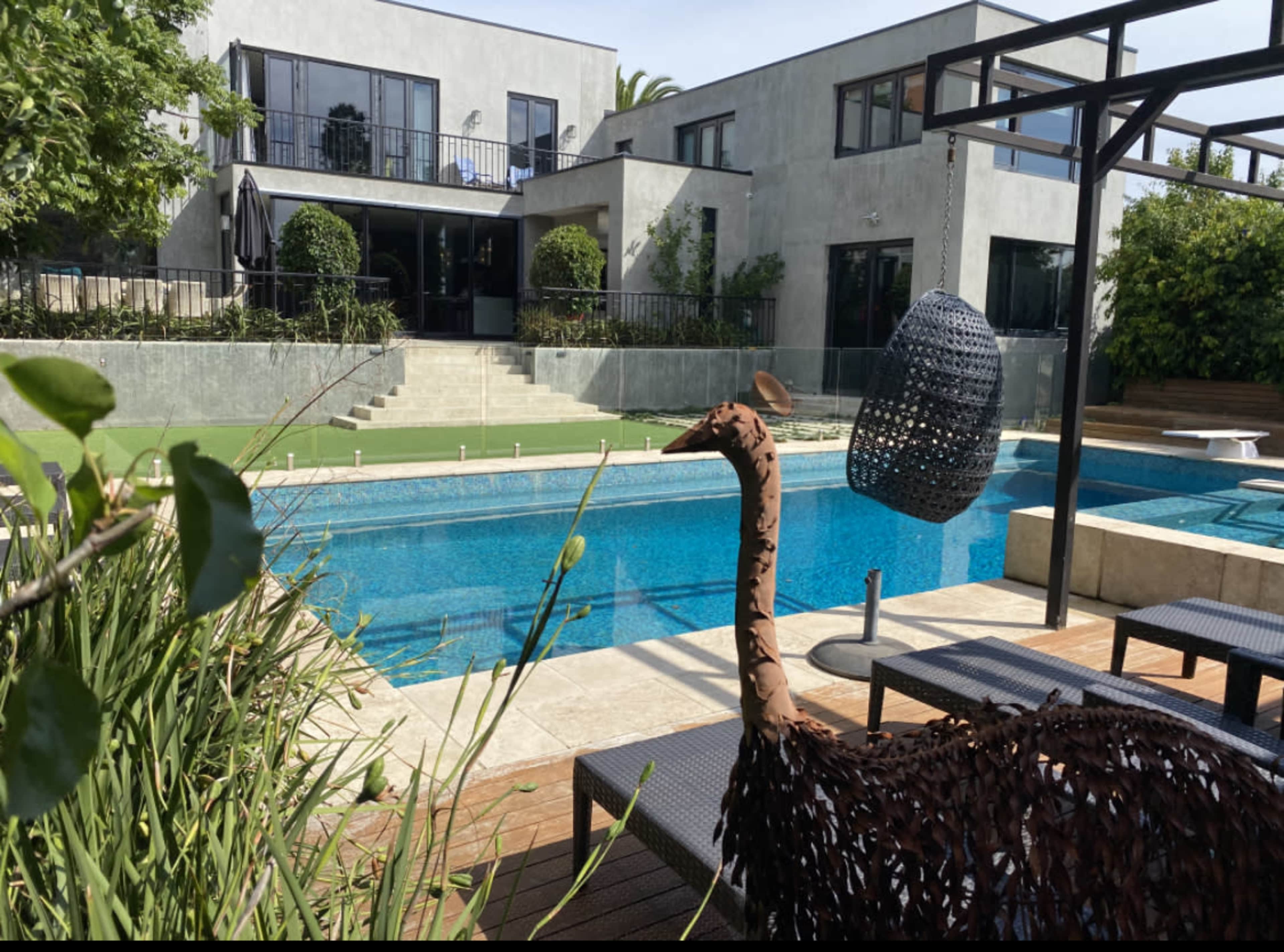 A metal sculpture of an ostrich stands near a swimming pool, with a modern house and landscaped greenery in the background.
