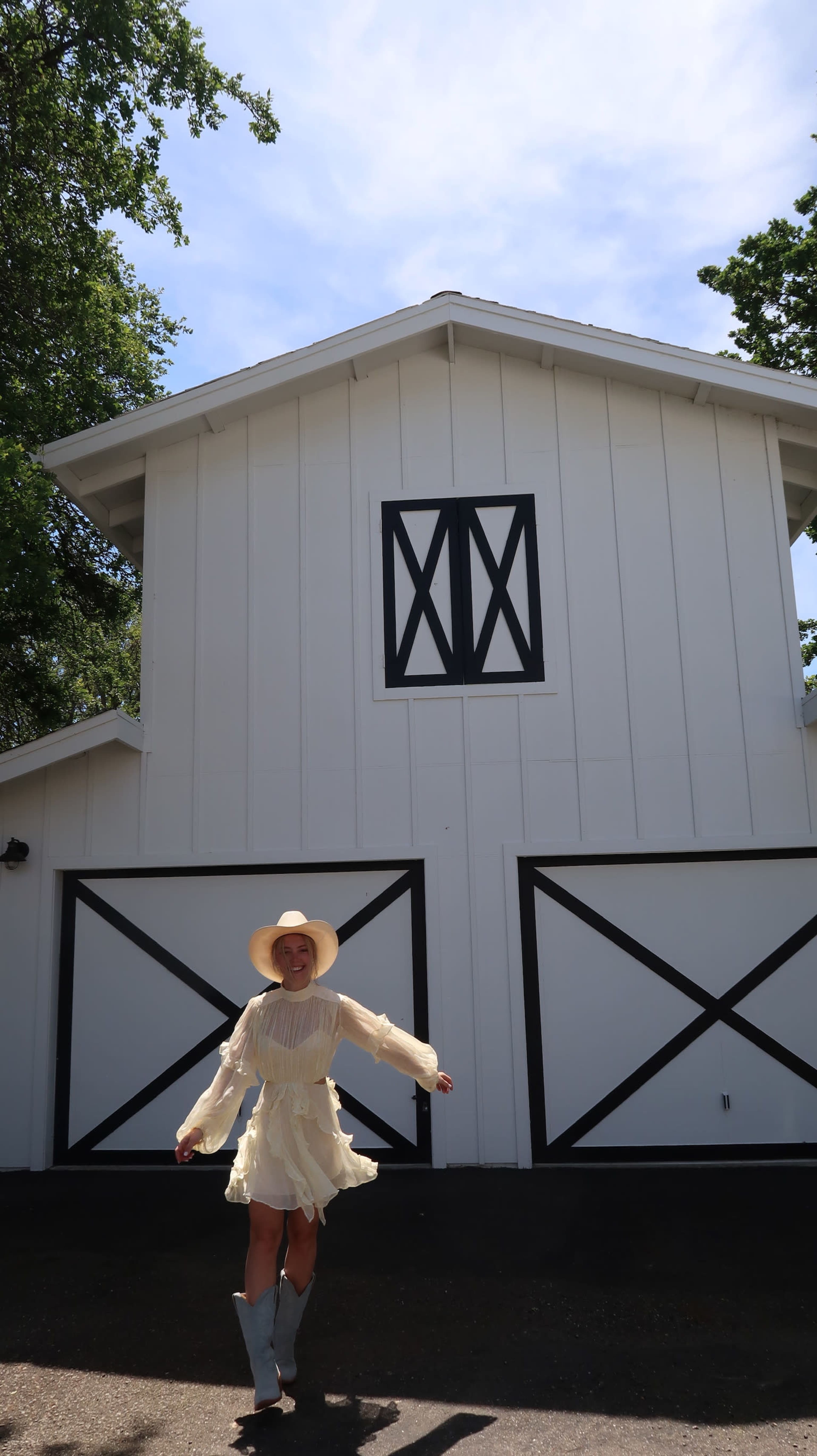 A person wearing a light dress and a wide-brimmed hat is standing in front of a white barn with black doors and windows.
