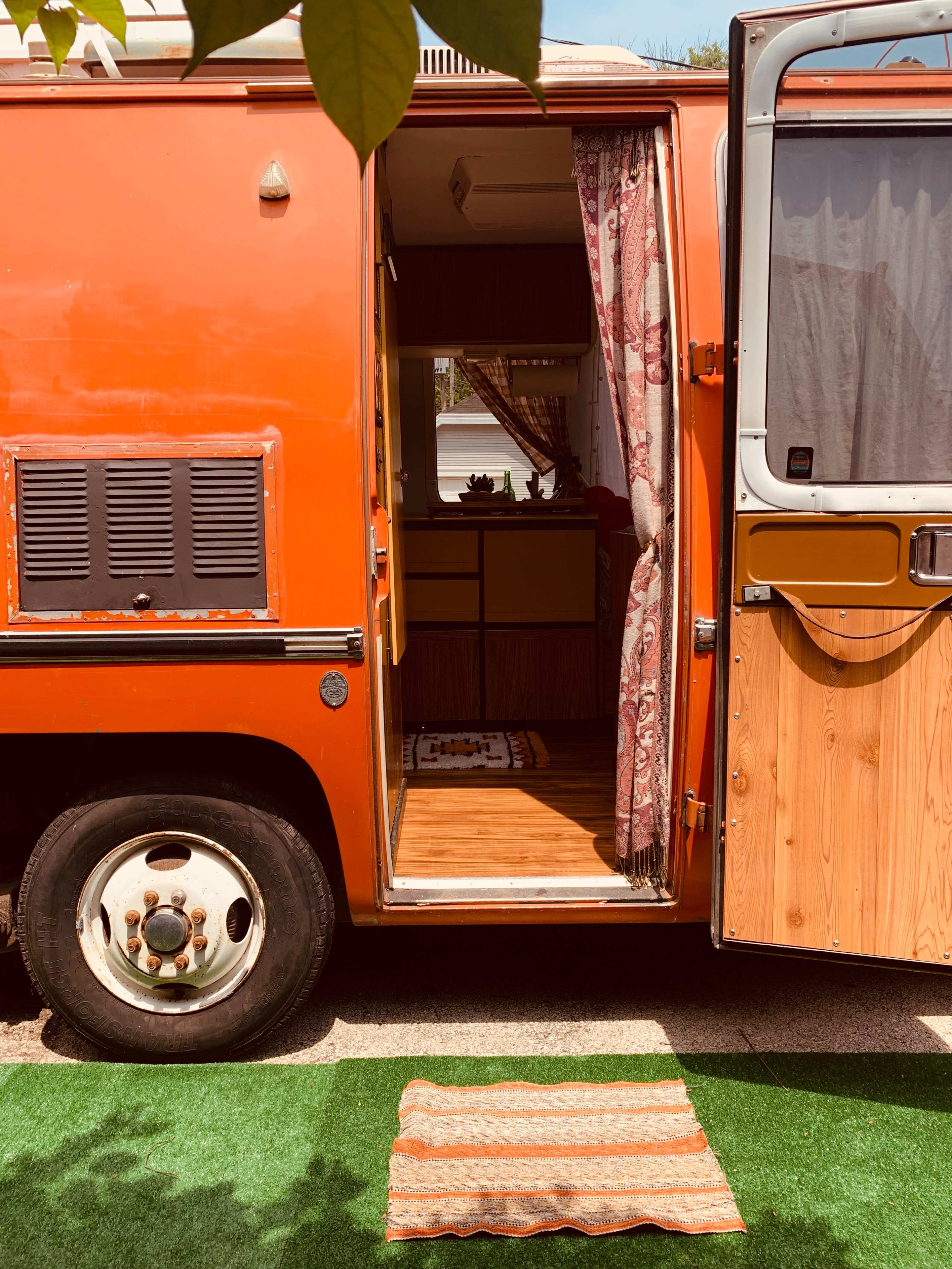 An orange vintage camper van stands with its door open, revealing a wooden interior and a welcome mat on the ground.