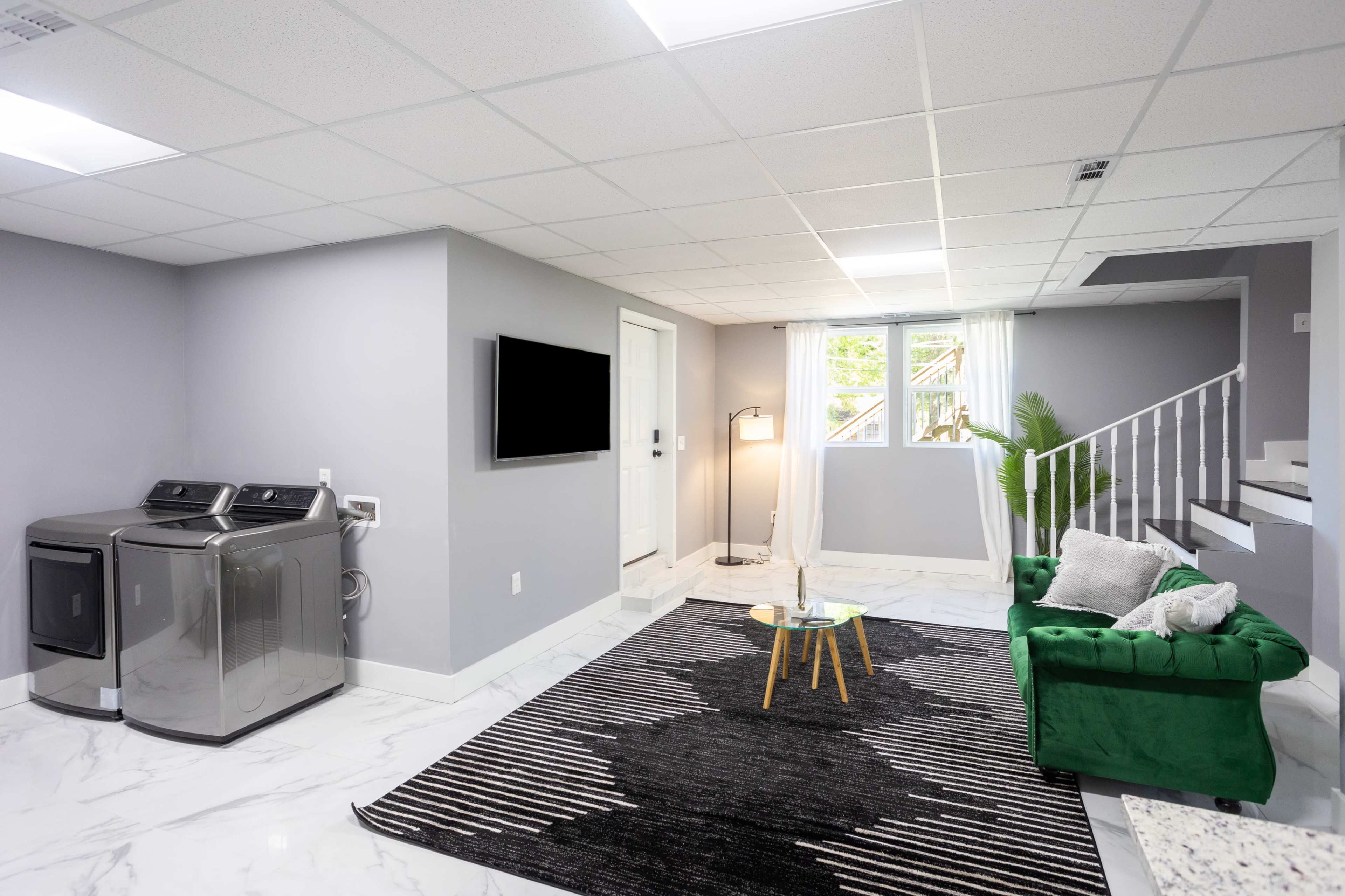 A modern basement features a laundry area, a television, a green sofa, and a staircase, with marble flooring and a striped rug.