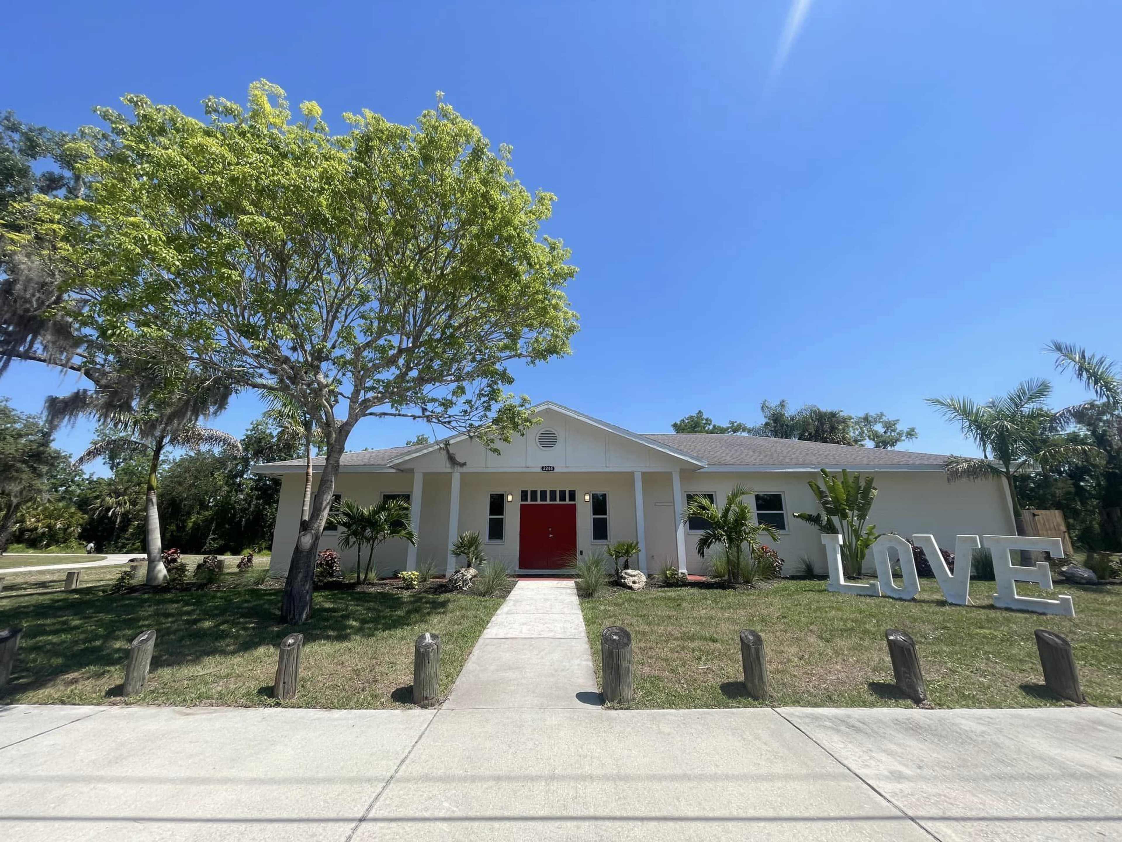 A single-story house with a red front door and large "LOVE" letters in the front yard is surrounded by greenery under a clear blue sky.