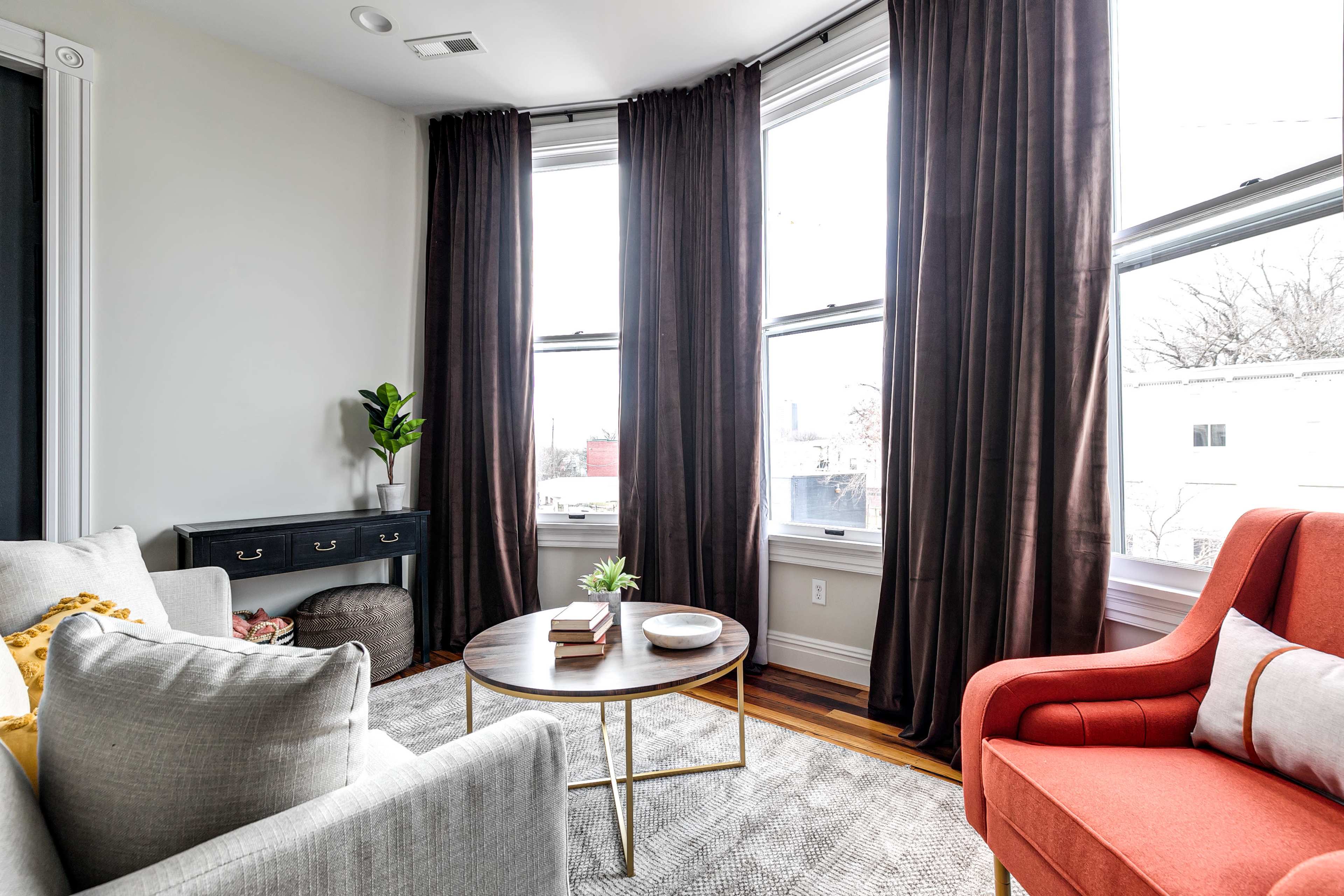 A cozy living room with a gray couch, a red armchair, a round coffee table, and large windows adorned with dark curtains.