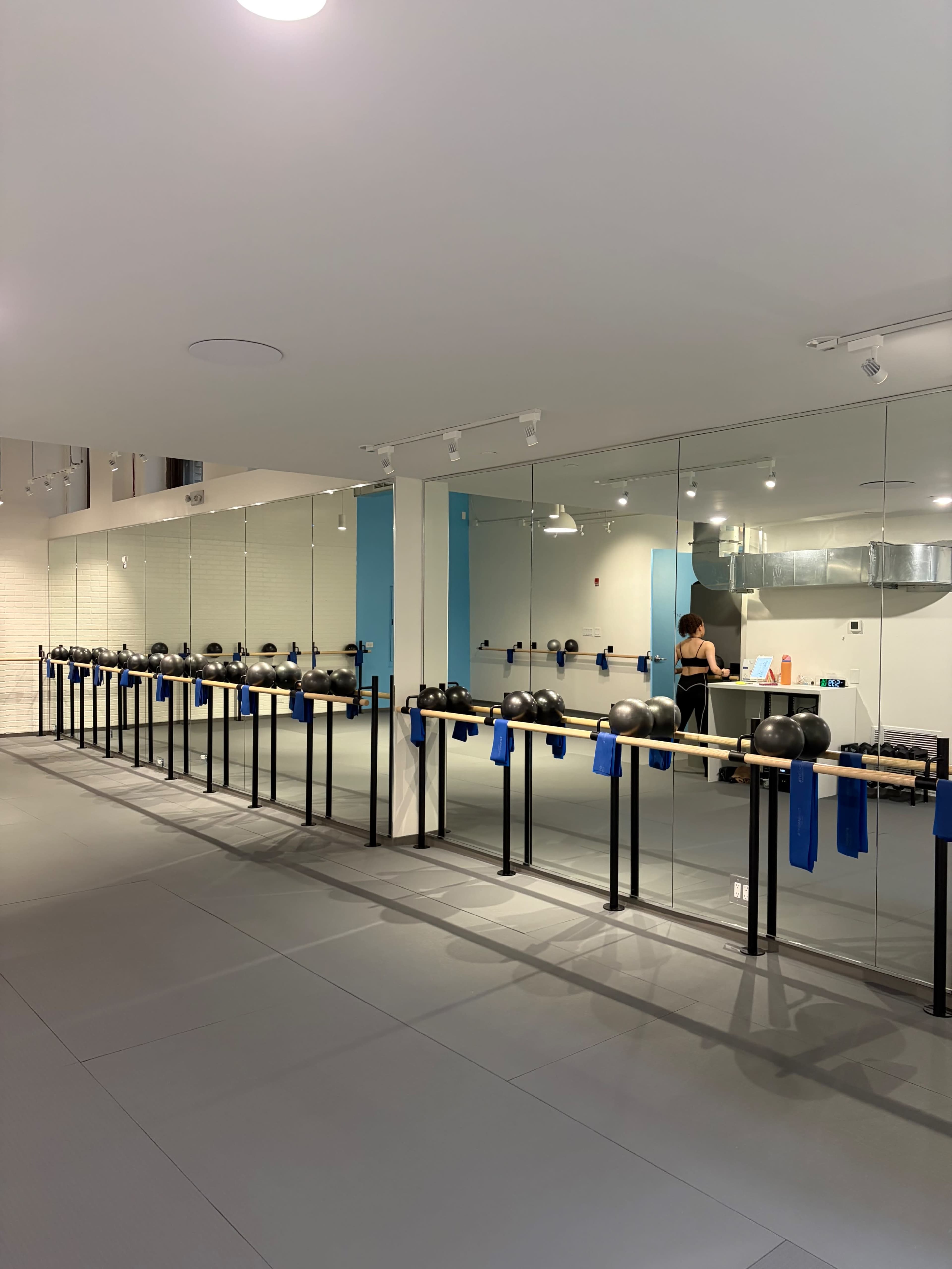 The image shows a mirrored fitness studio with a row of weights on stands and a person exercising in the background.
