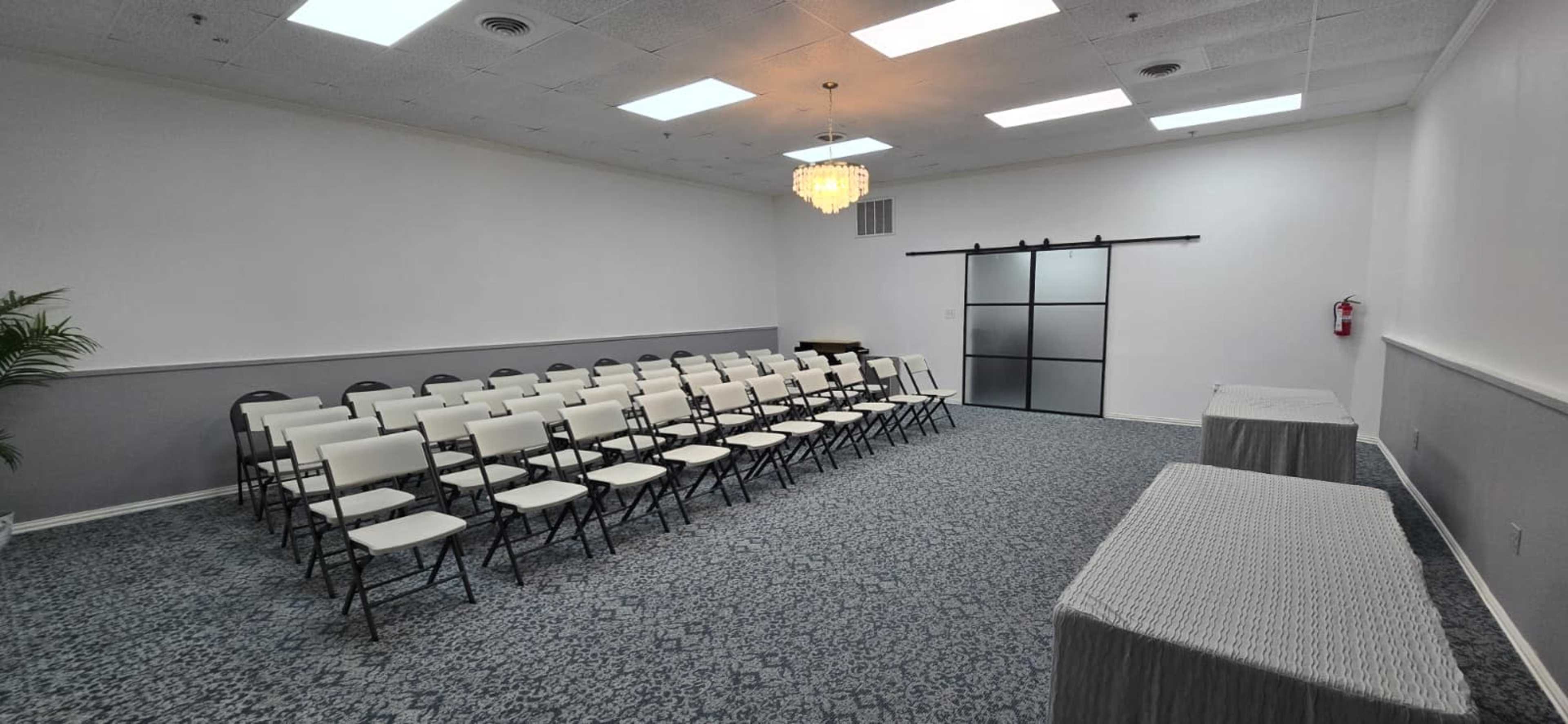 The image shows a vacant conference room set up with rows of white folding chairs facing a table near a sliding glass door, with a chandelier overhead.