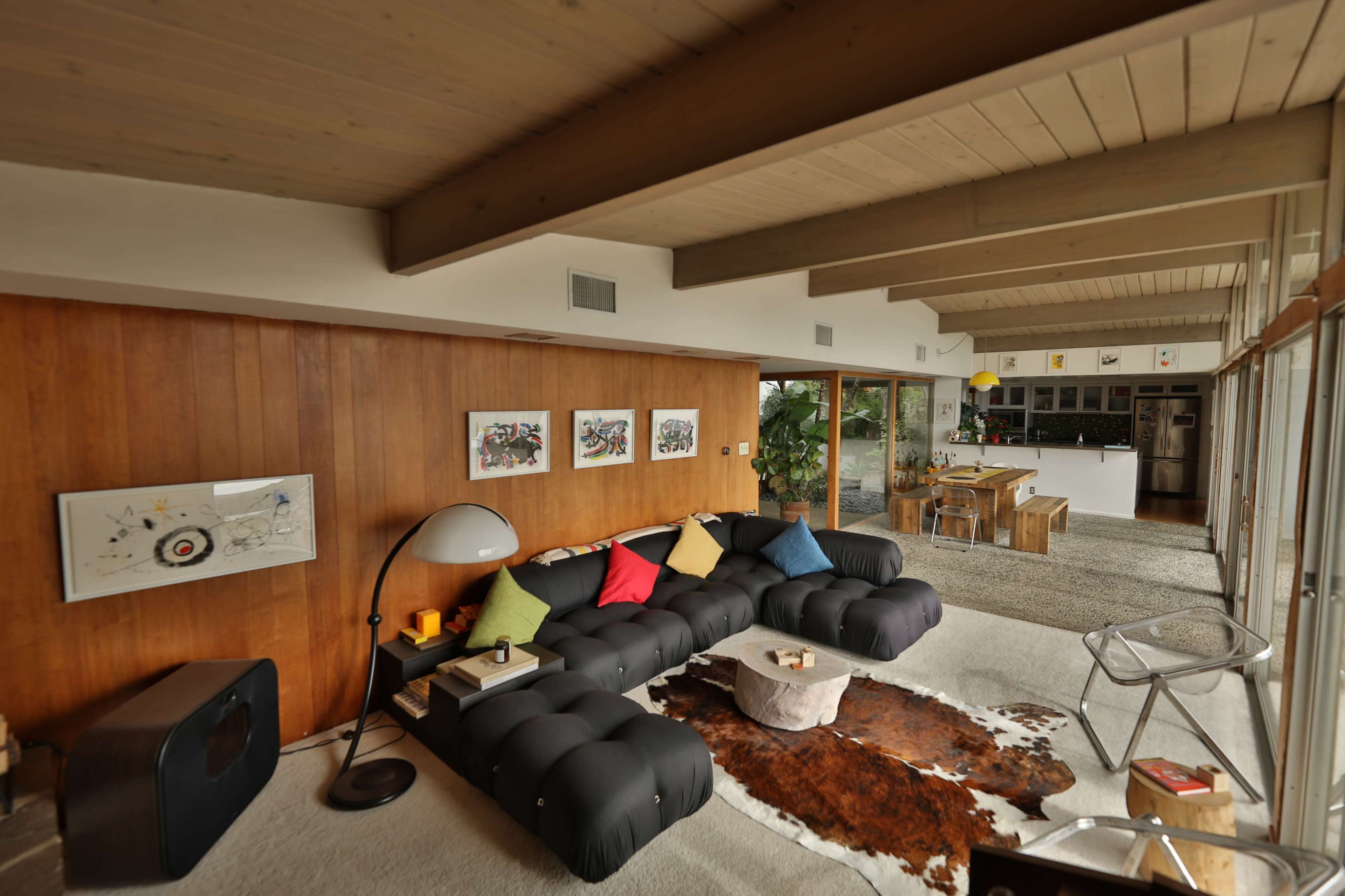 The image shows a modern living room with a black sectional sofa, colorful cushions, wooden walls, and an open kitchen in the background.