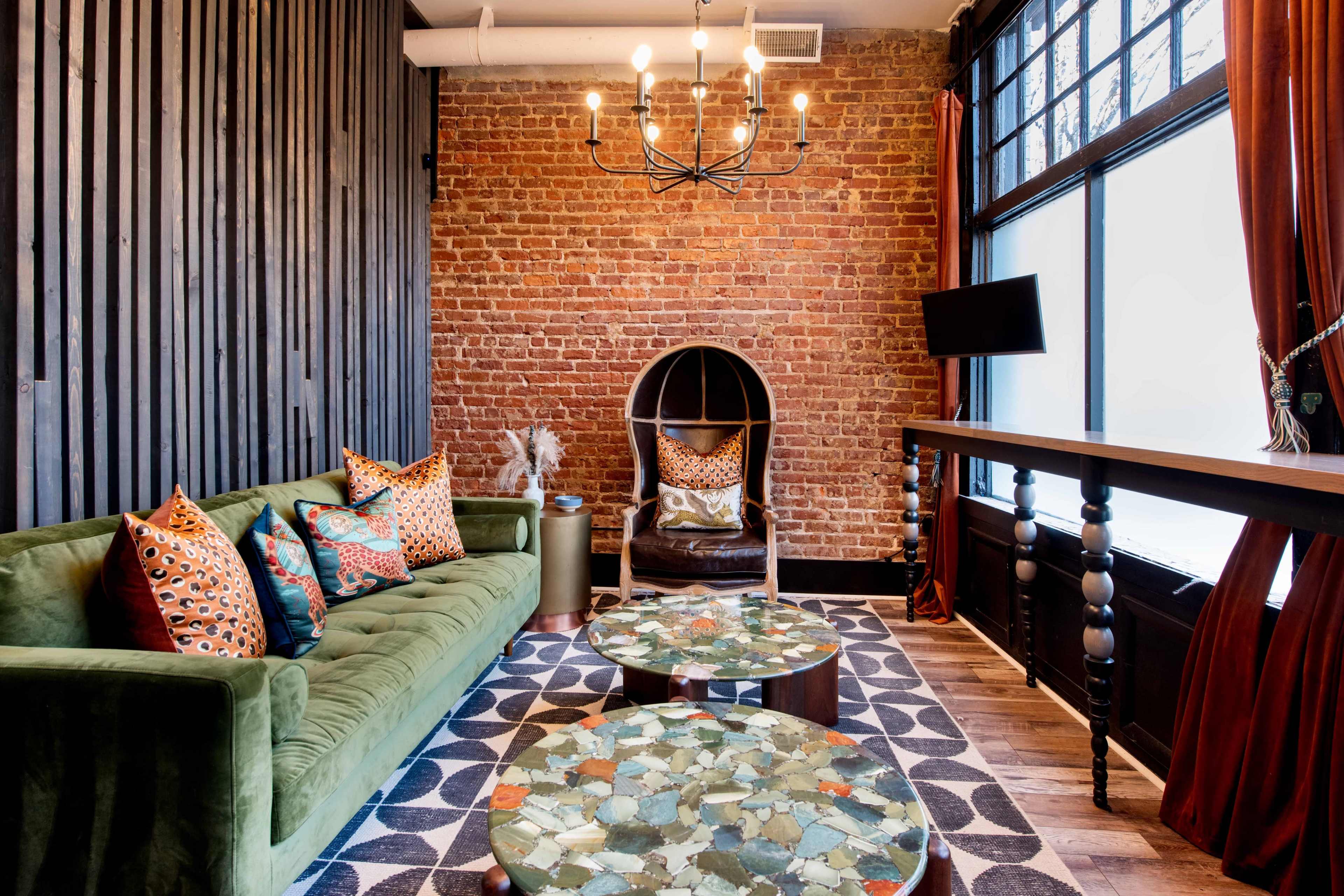 A cozy lounge area featuring a green sofa with patterned cushions, two circular coffee tables, and a brick wall backdrop.