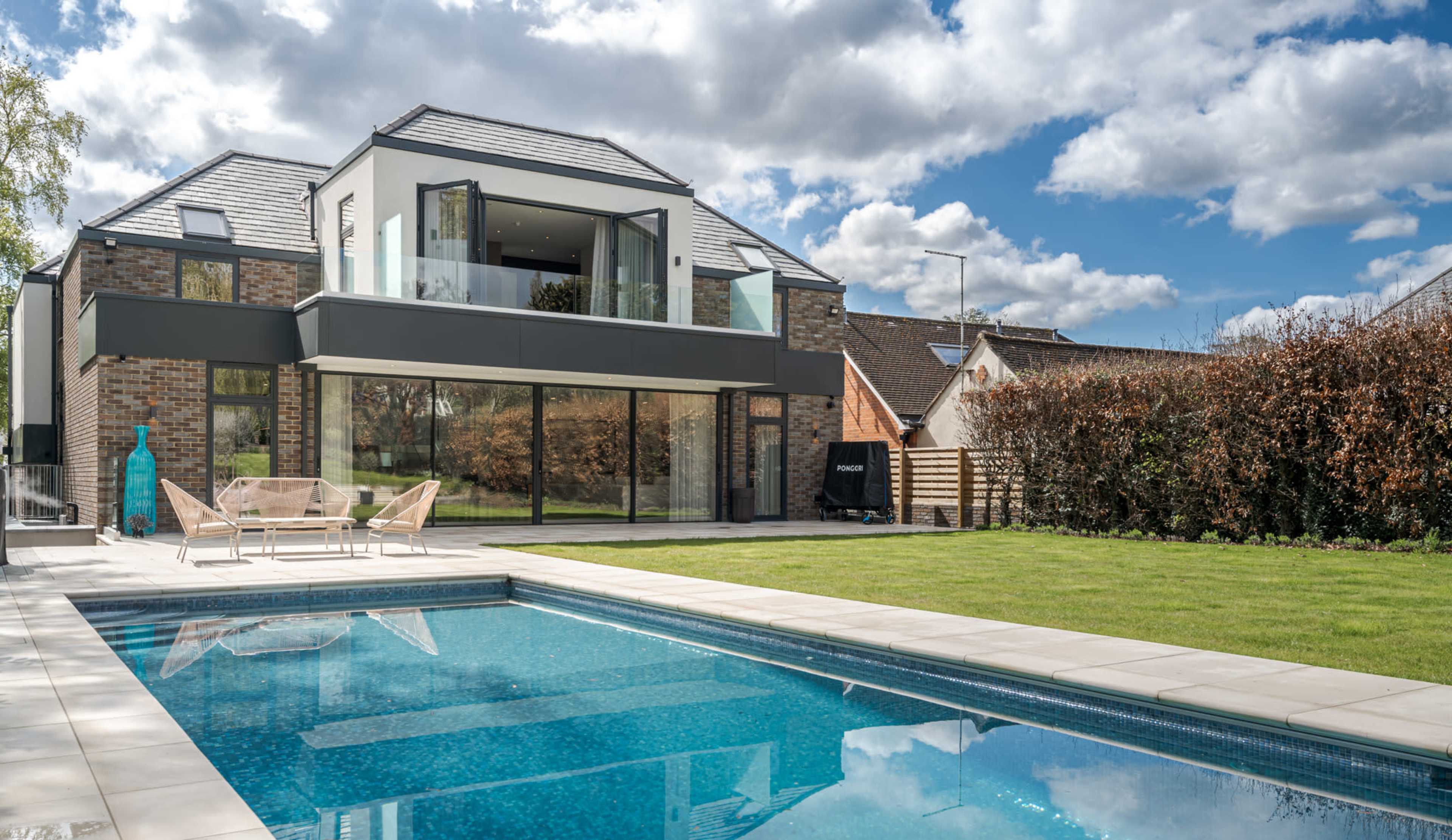 A modern two-story house with large windows overlooks a swimming pool and green lawn under a partly cloudy sky.