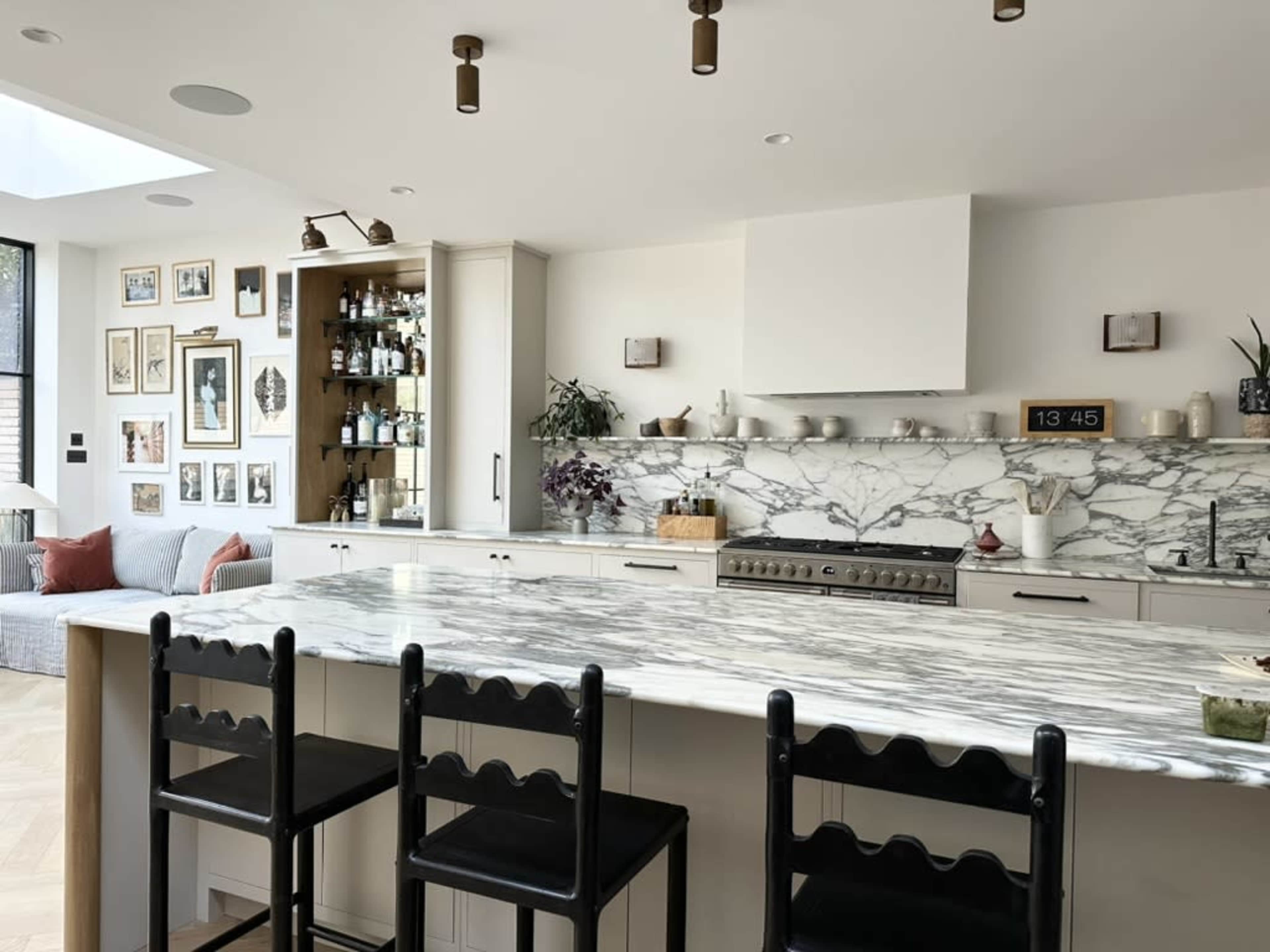 The image shows a modern kitchen featuring a marble countertop with three black bar stools, a stove, and shelves with various bottles and decorative items on the wall.