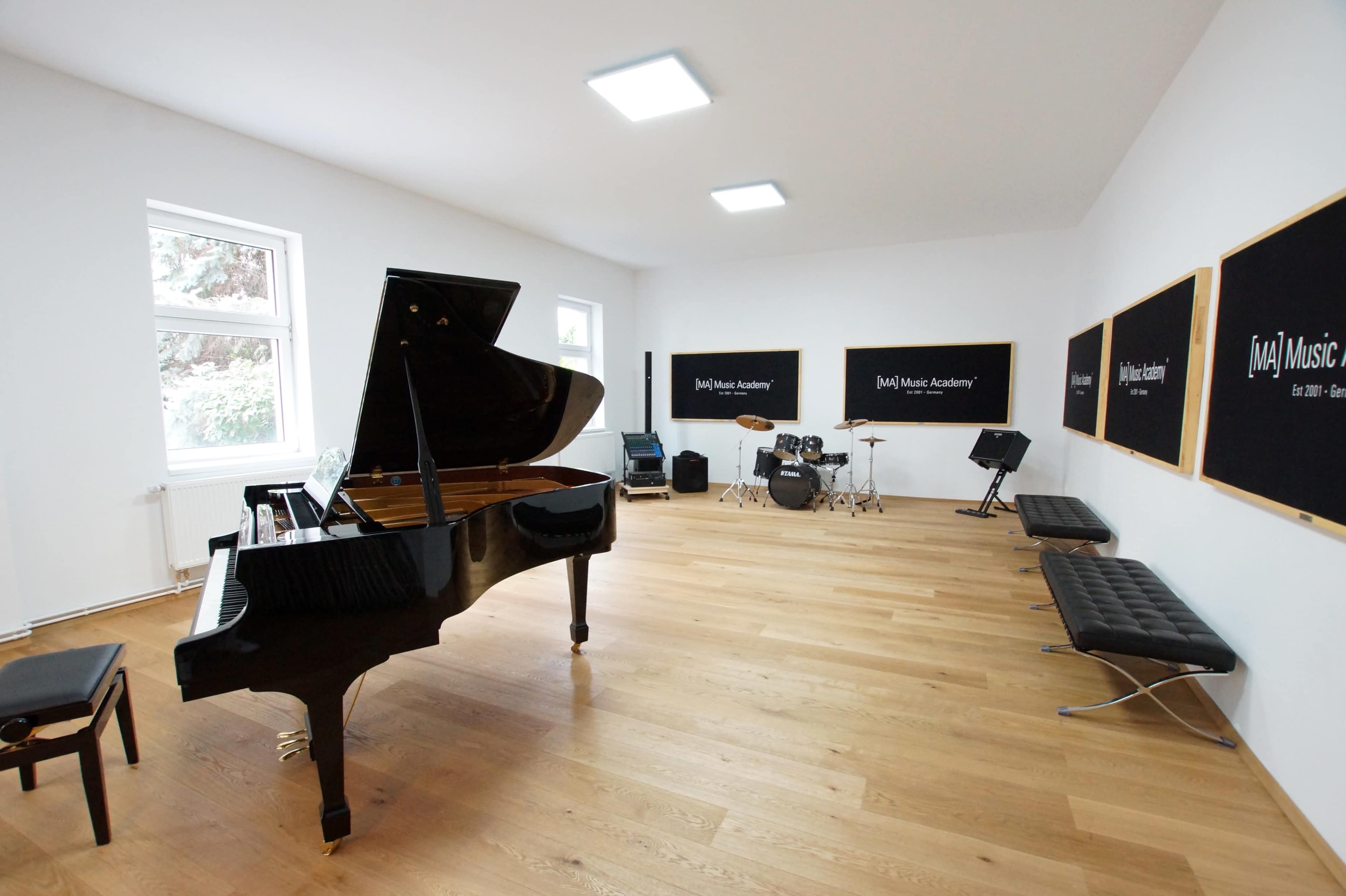 The image shows a music studio with a grand piano, a set of drums, and several benches arranged against white walls adorned with black signs.