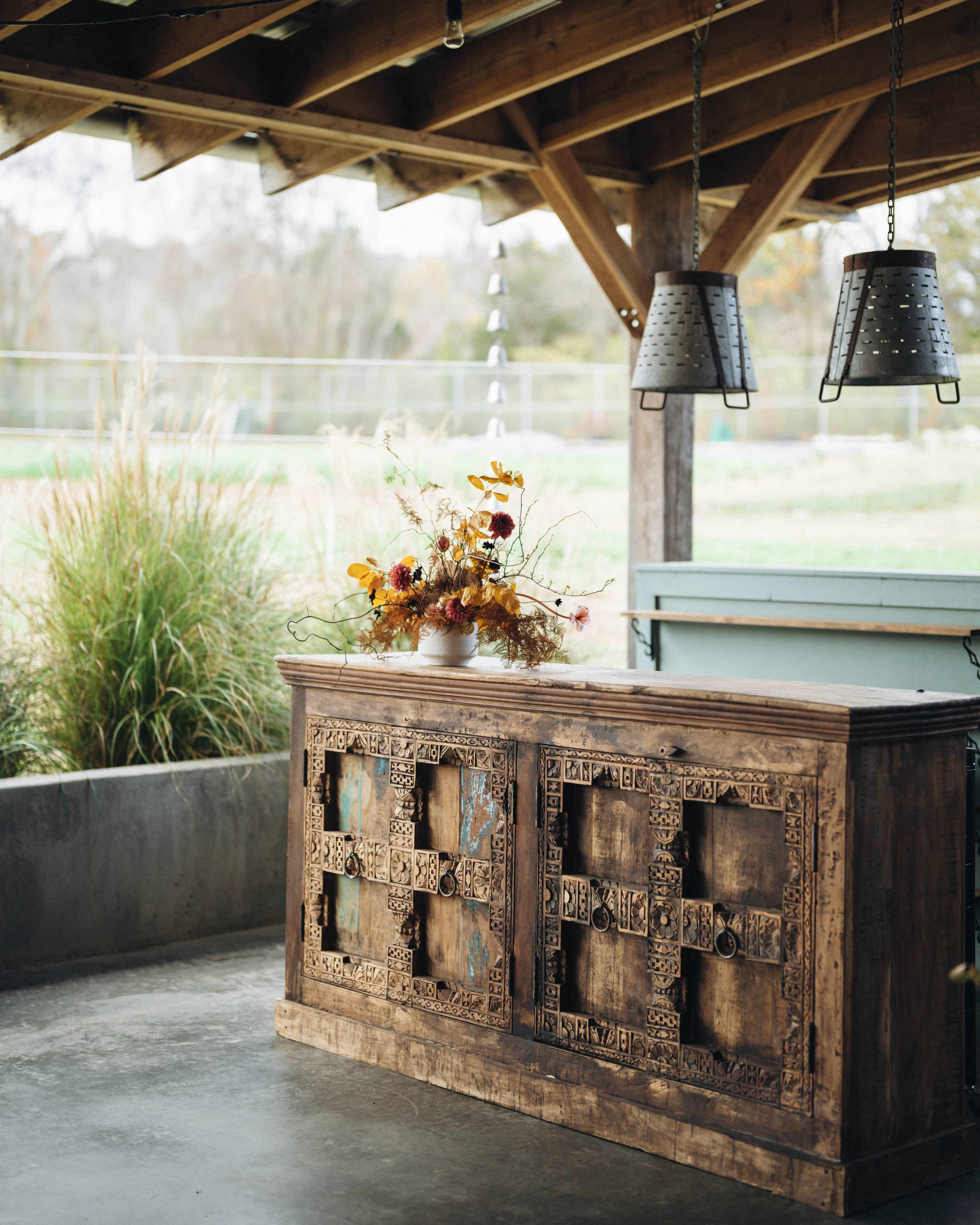 A wooden sideboard with intricate carvings is placed under a covered outdoor space, adorned with a vase of flowers and hanging metal lamps above.