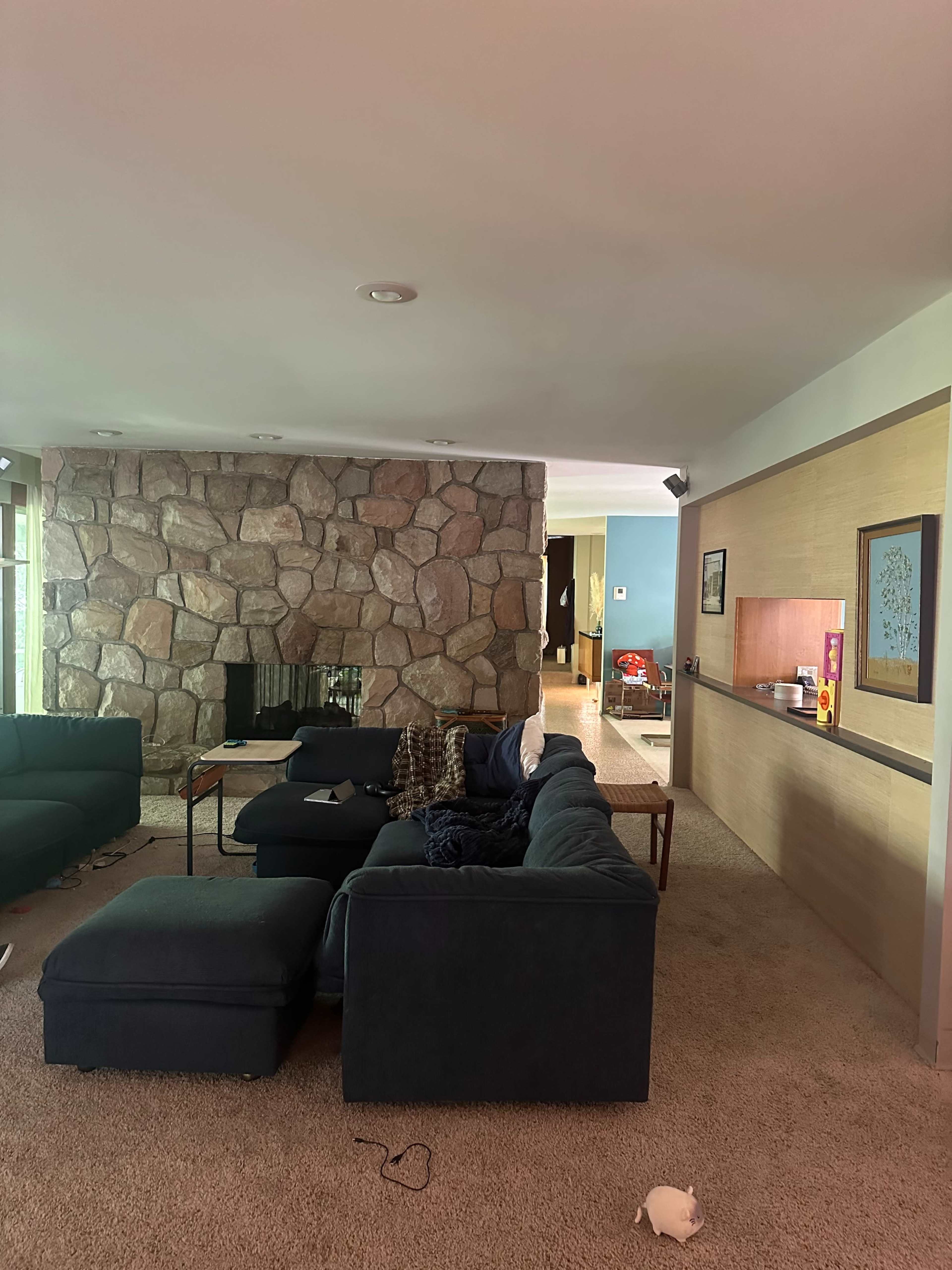 The image shows a living room with a stone fireplace, a dark blue sectional sofa, and a carpeted floor, leading to a hallway and another room.