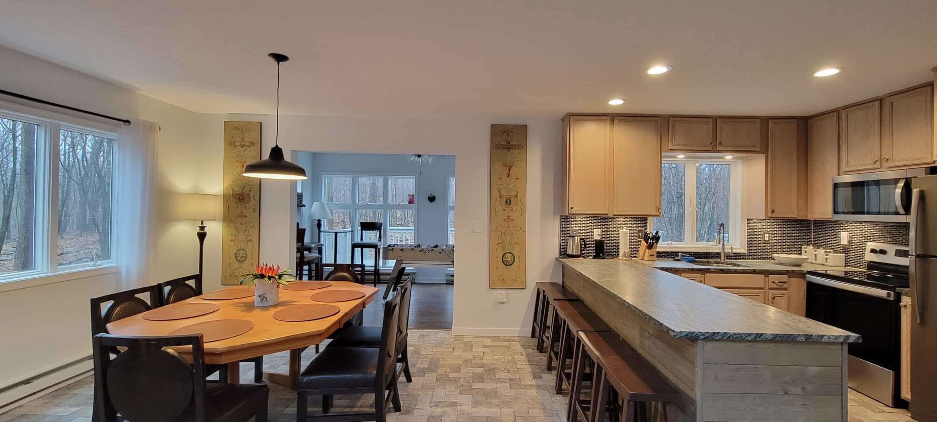 The image shows a modern kitchen and dining area with a round wooden table, chairs, and a long counter with stools, all set in a well-lit space with large windows facing a wooded area.