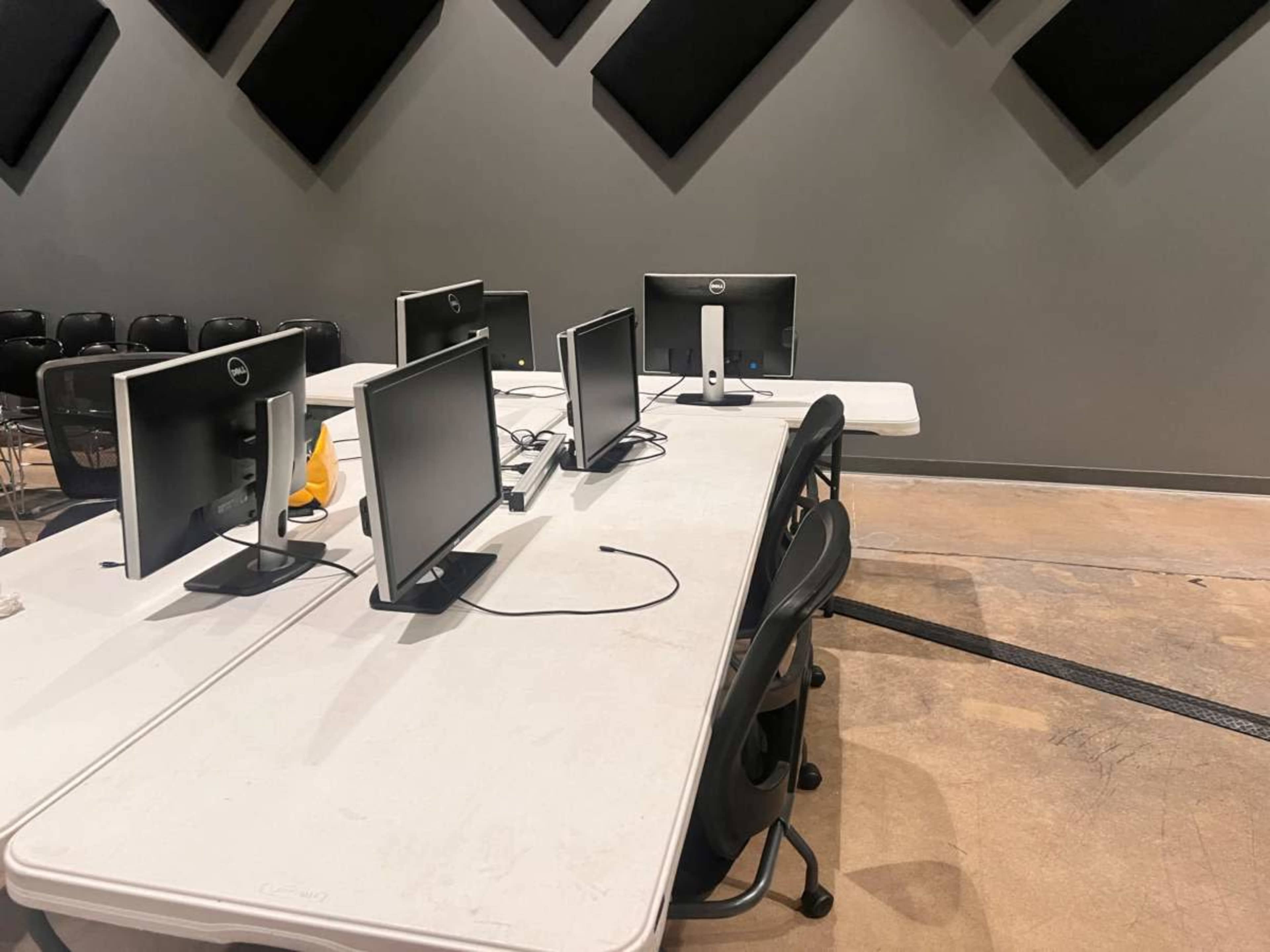 A row of four computer monitors is arranged on a table in a room with soundproofing panels on the walls.