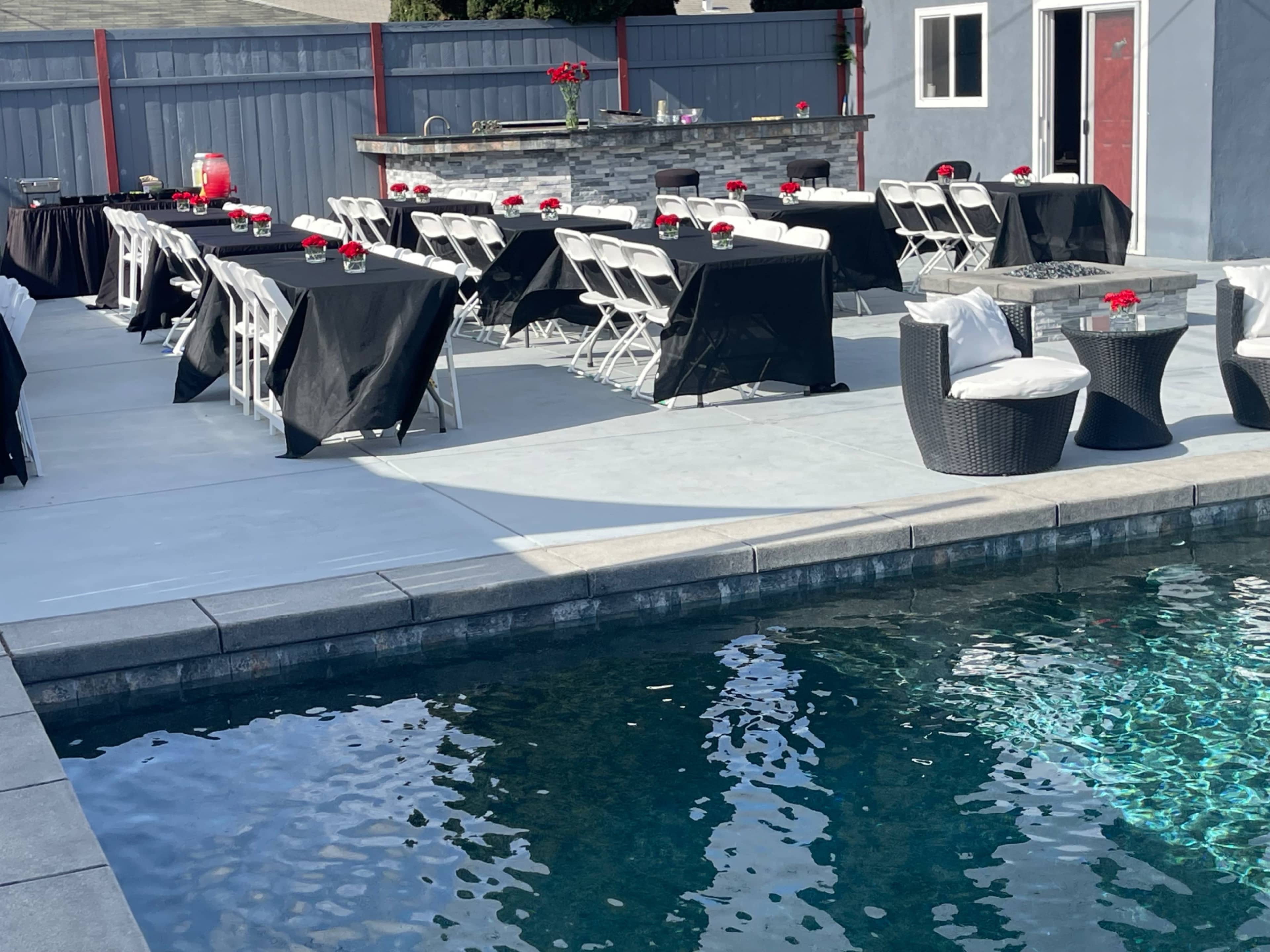 The image shows a backyard setting with neatly arranged tables covered in black tablecloths, a swimming pool, and a bar area with red accents.